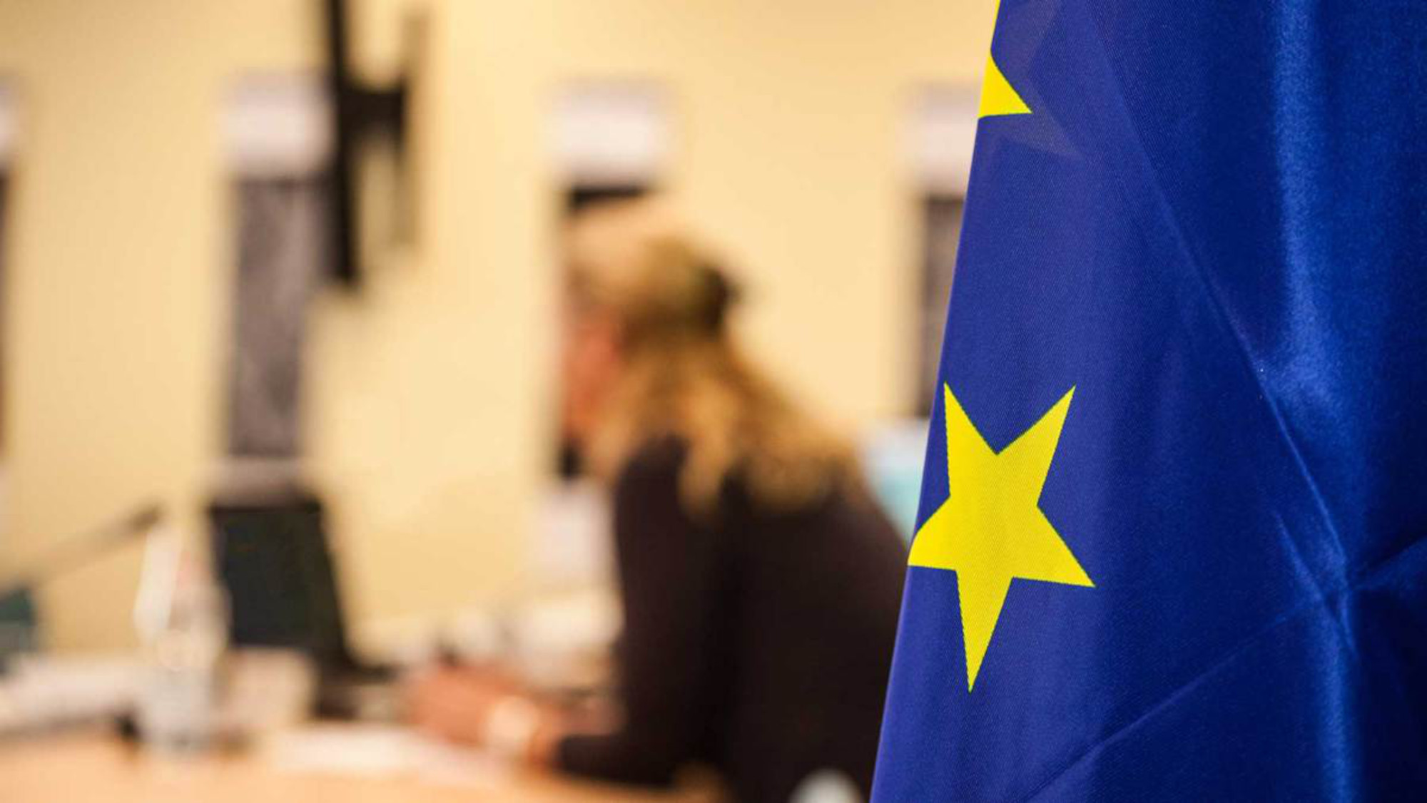 A part of the EU flag is focused in the foreground, in the background a person is sitting at a desk and working on a laptop.