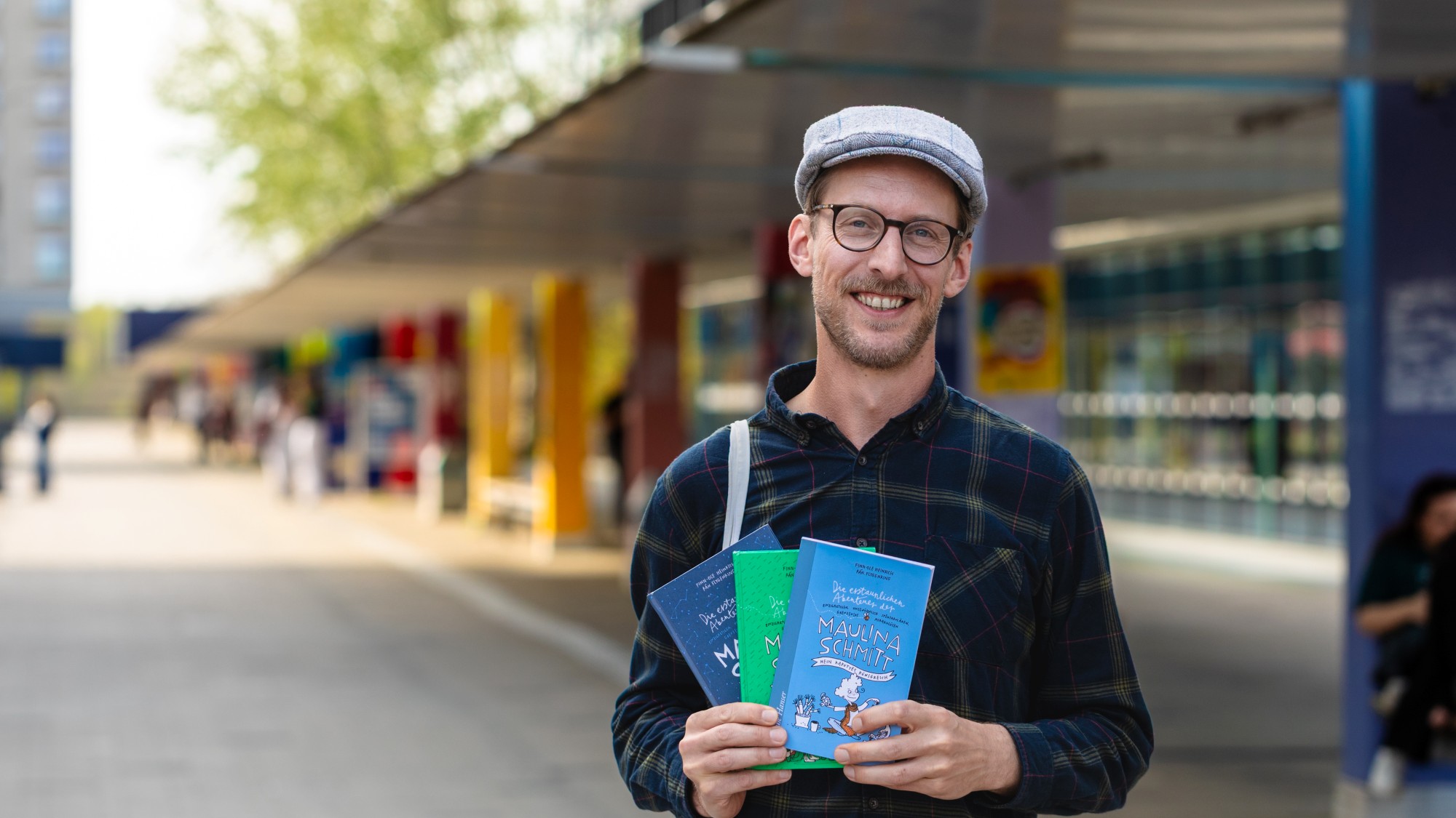 Ein Mann mit Schiebermütze und Brille lächelt in die Kamera und hält Bücher in den Händen.
