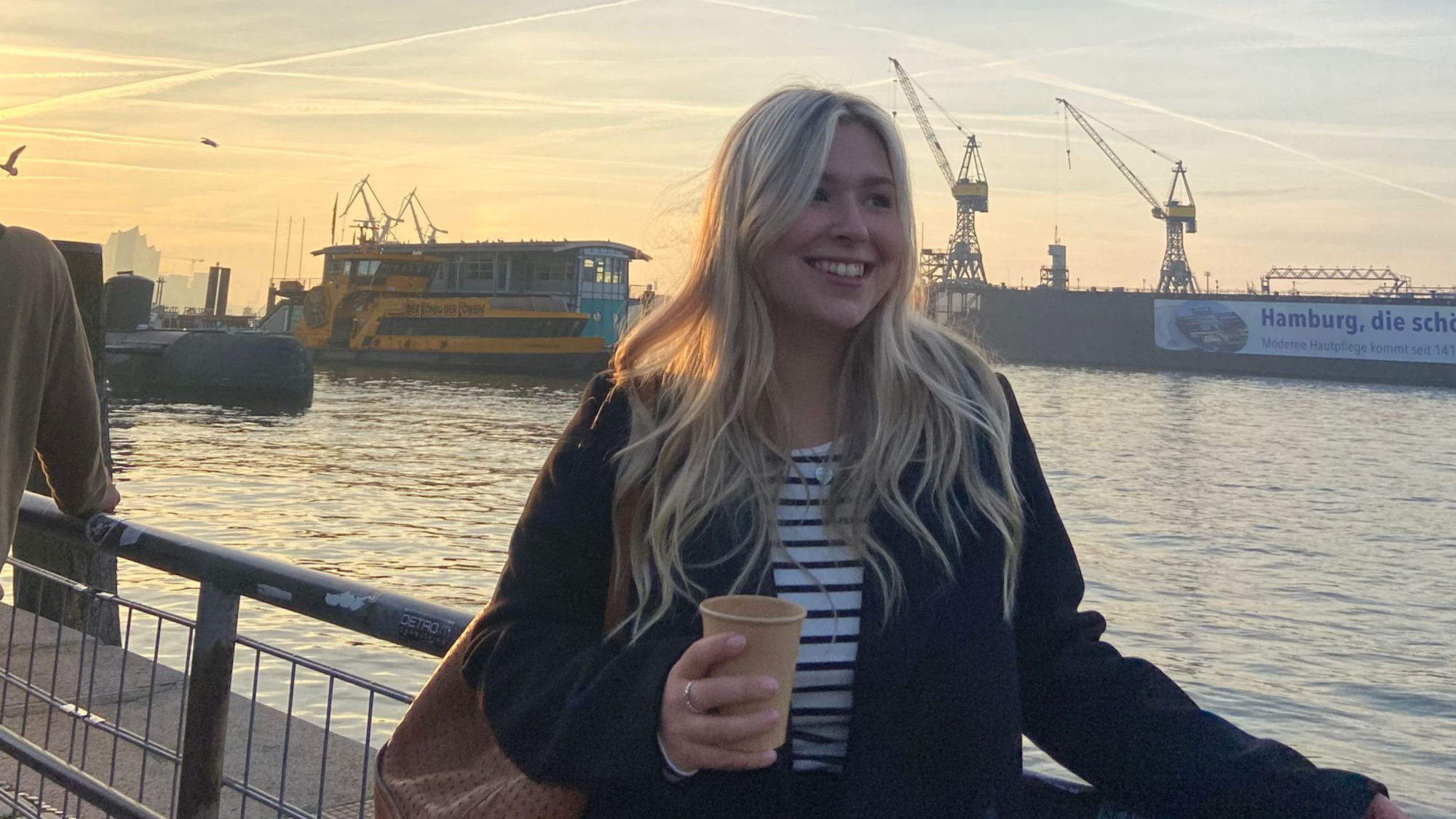 Emily is standing in front of the Elbe in Hamburg. She is holding a coffee cup in her hand.