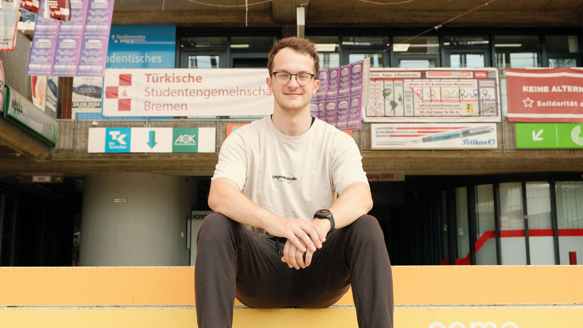 Florian sitzt auf der Treppe in der Glashalle und lächelt in die Kamera.
