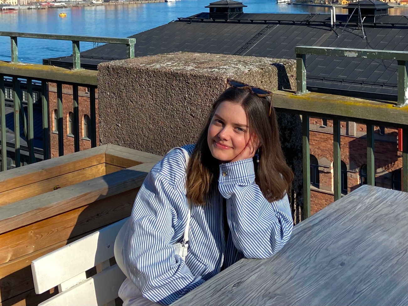 Hannah is sitting on a bench in front of a lake in a blue and white striped jacket.