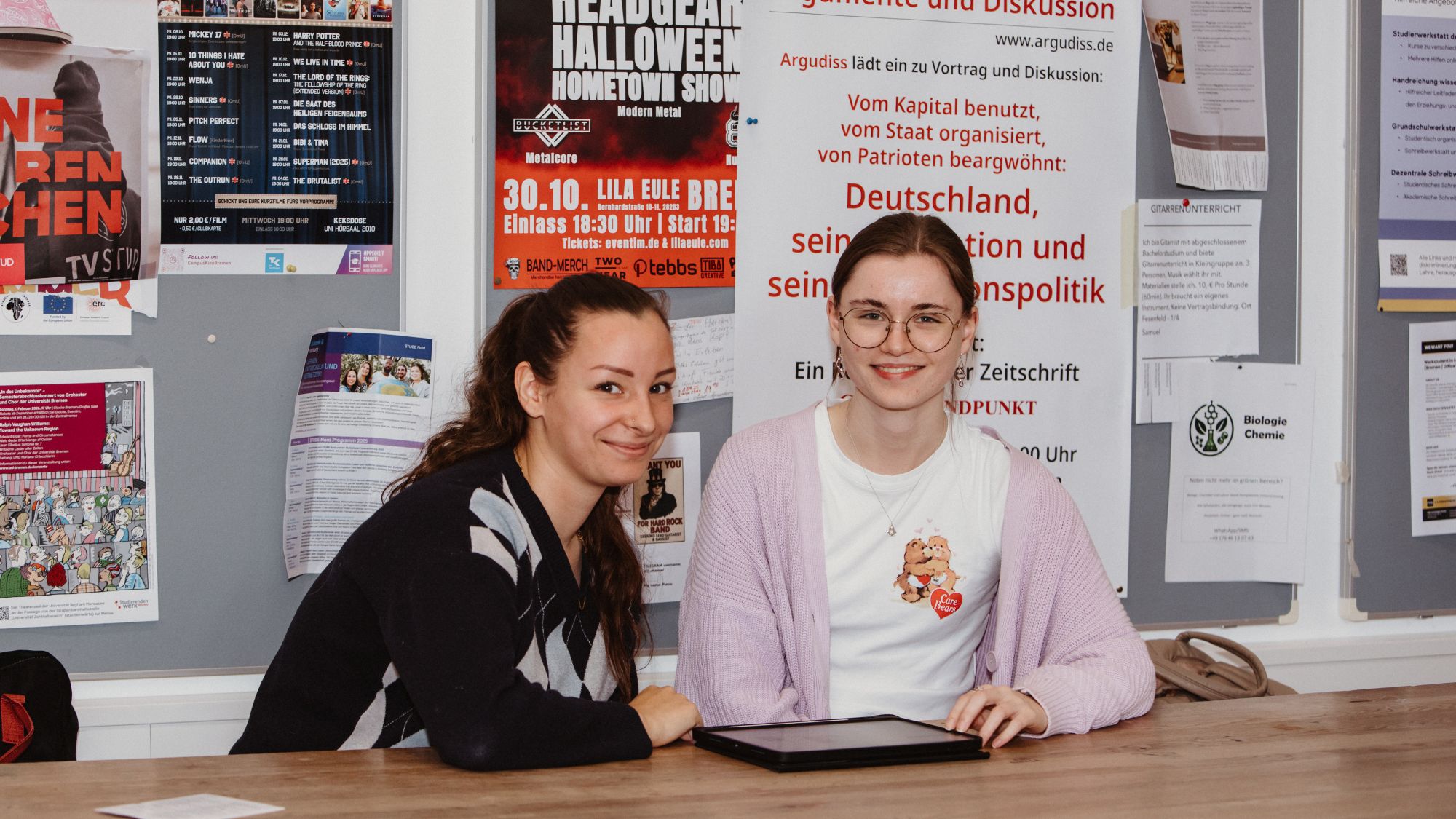 Zwei Personen sitzen mit einem Tablet an einem Tisch und schauen in die Kamera