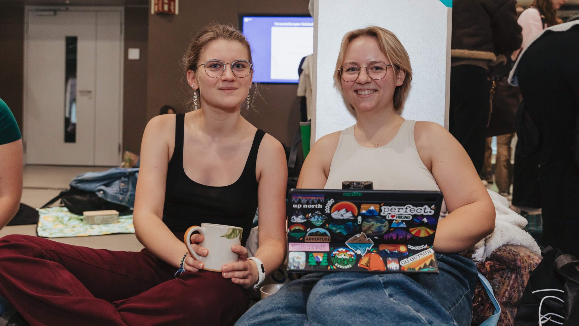 Zwei Frauen sitzen zusammen auf dem Boden. Die eine hält eine Tasse in der Hand und die andere hat ein Laptop auf dem Schoss.