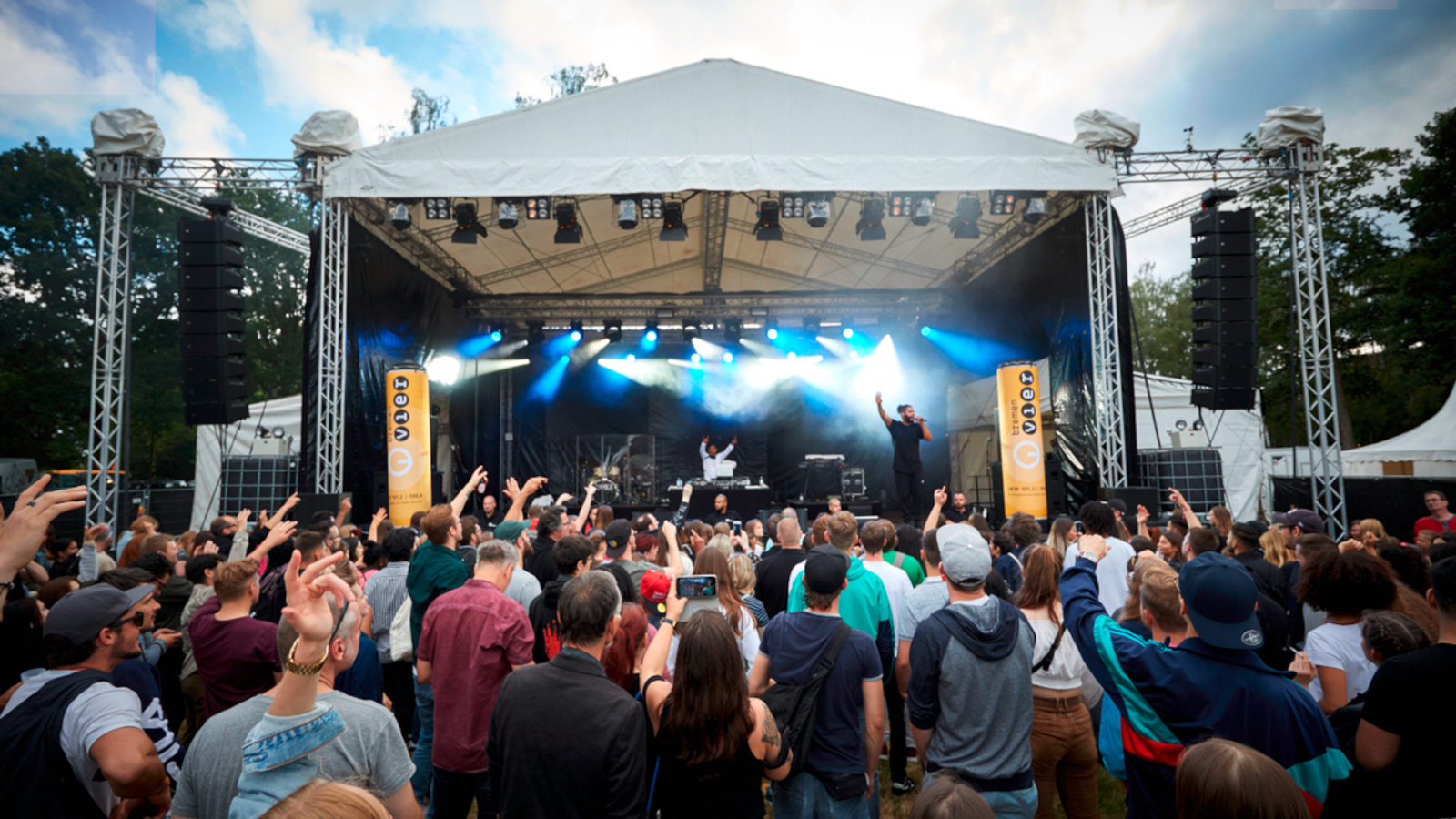 Ein Künstler tritt auf einer Open Air Bühne auf. Davor steht eine Menge junger fröhlicher feiernder Menschen.