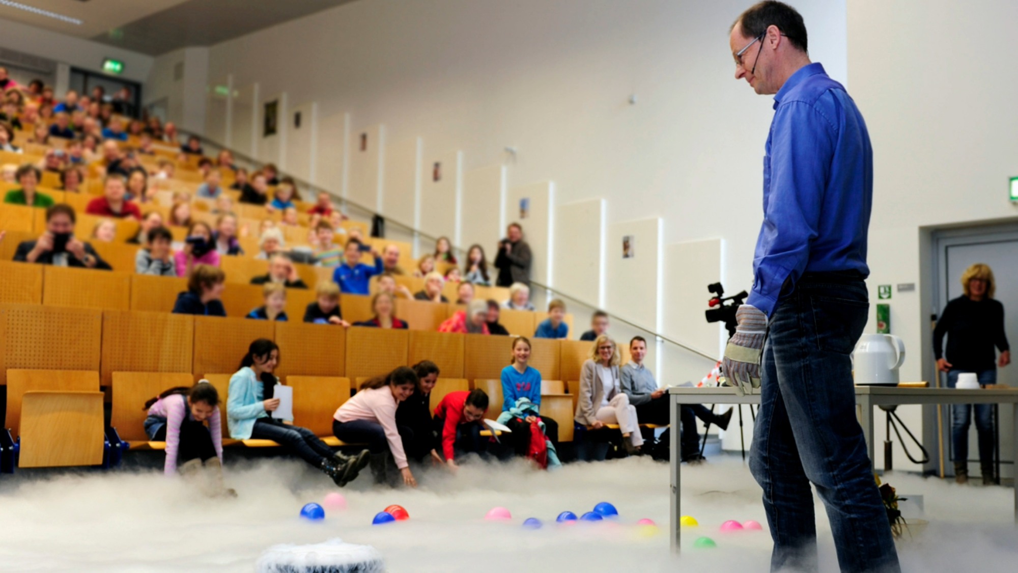 A professor stands at the front of the lecture hall and looks down at the fog on the floor. 