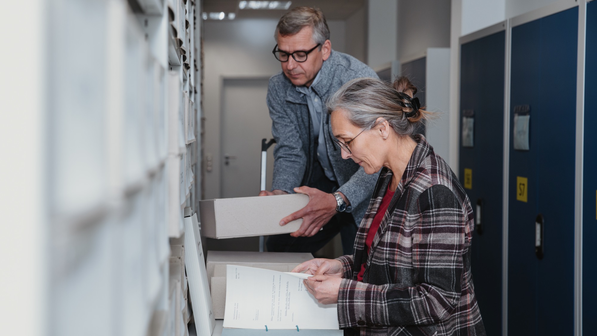 Two people are standing in front of a shelf looking at documents.