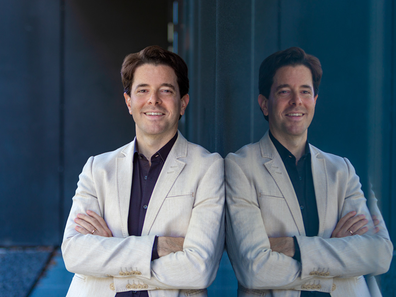 Mariano Chiacchiarini with folded arms in a white jacket leans against a pane of glass in which he is reflected.