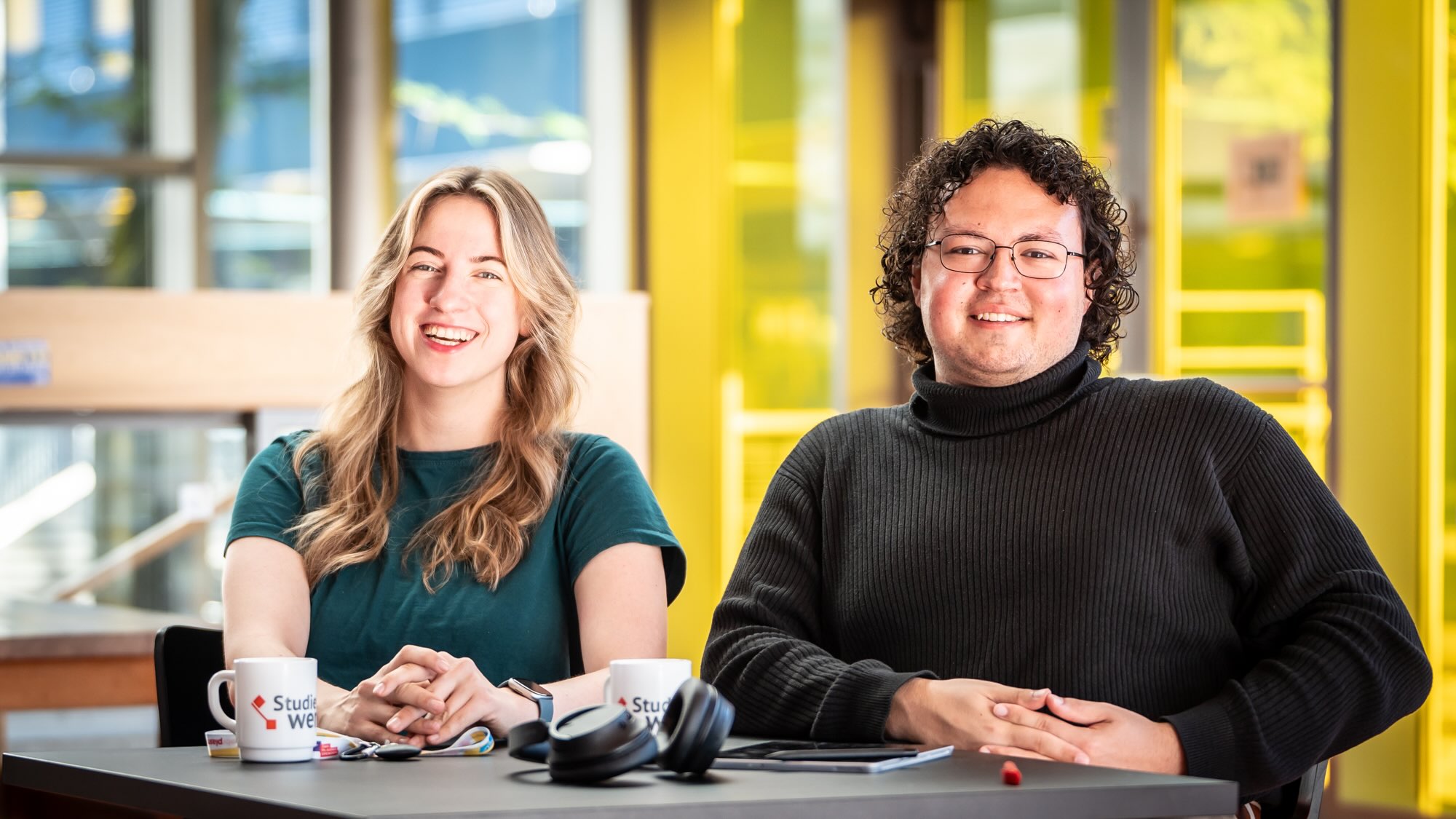 Paulina und Deniz sitzen nebeneinander an einem Tisch und lächeln in die Kamera.
