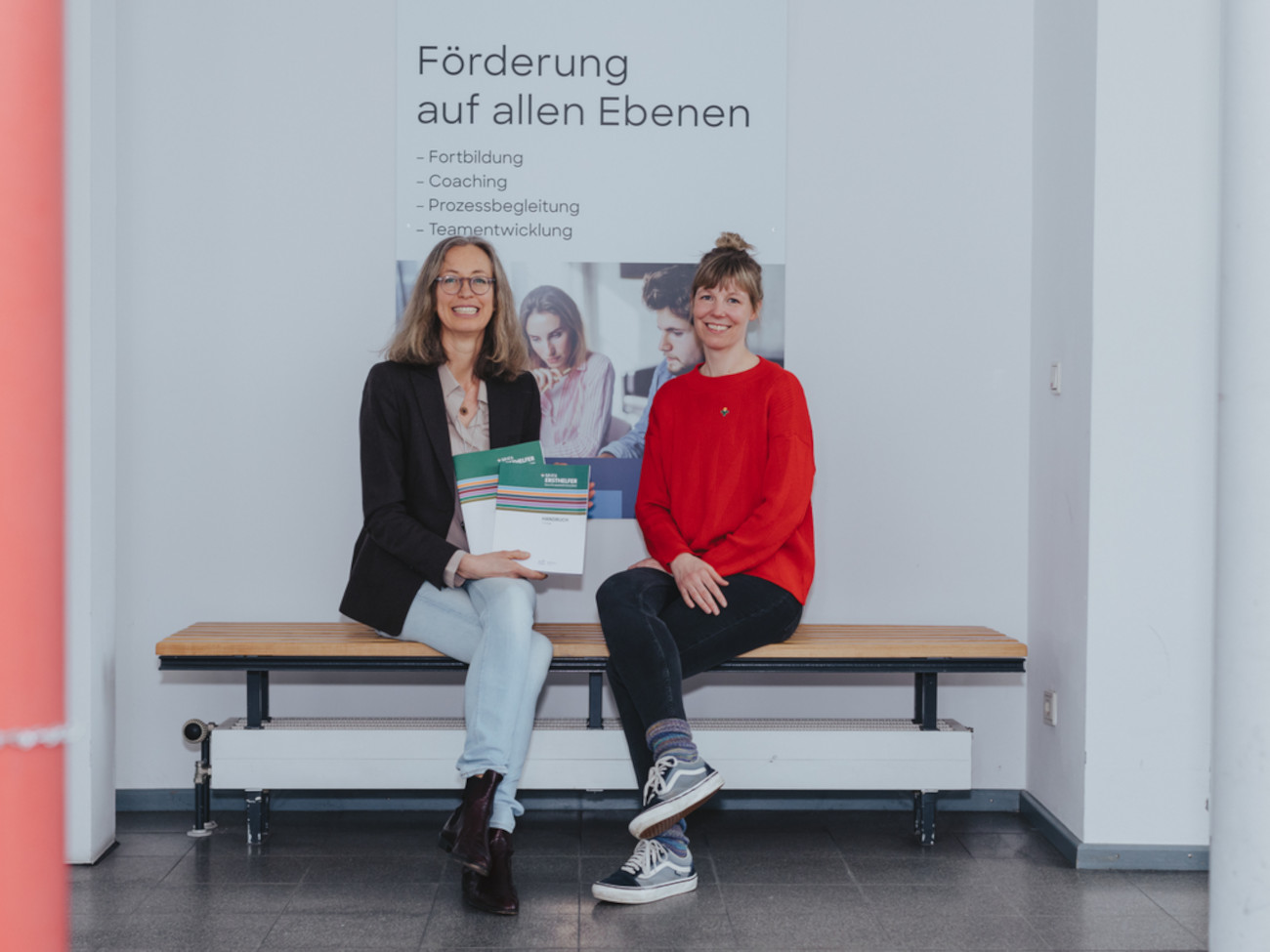 Simone Oelze und Alexandra Baumkötter sitzen auf einer Bank. Sie halten MHFA Kataloge in den Händen.