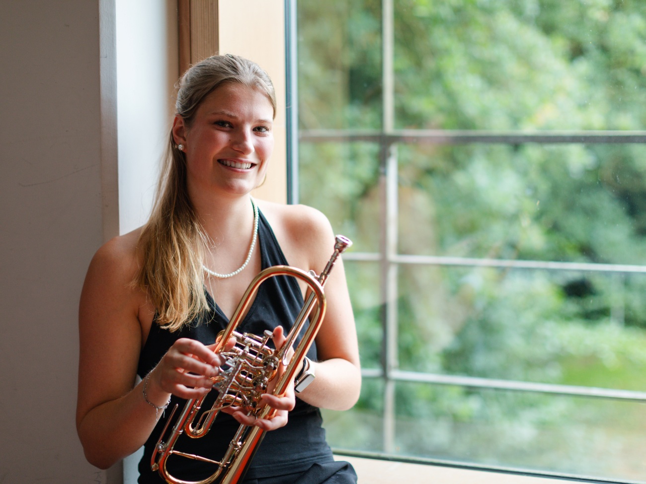 A woman with a trumpet in her hand is standing in front of a window.