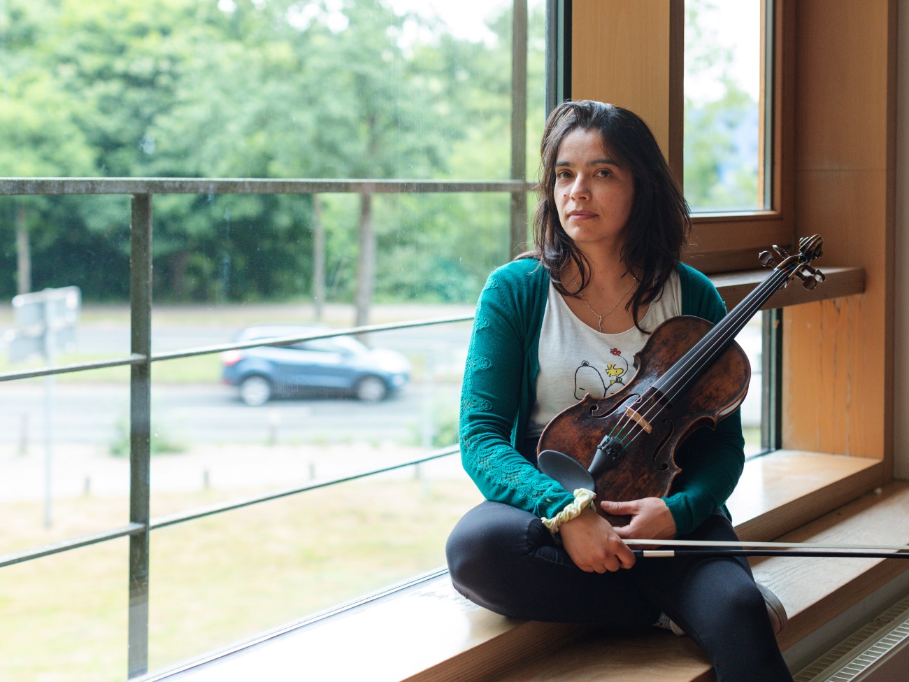 A woman is sitting on a windowsill with a viola on her lap.