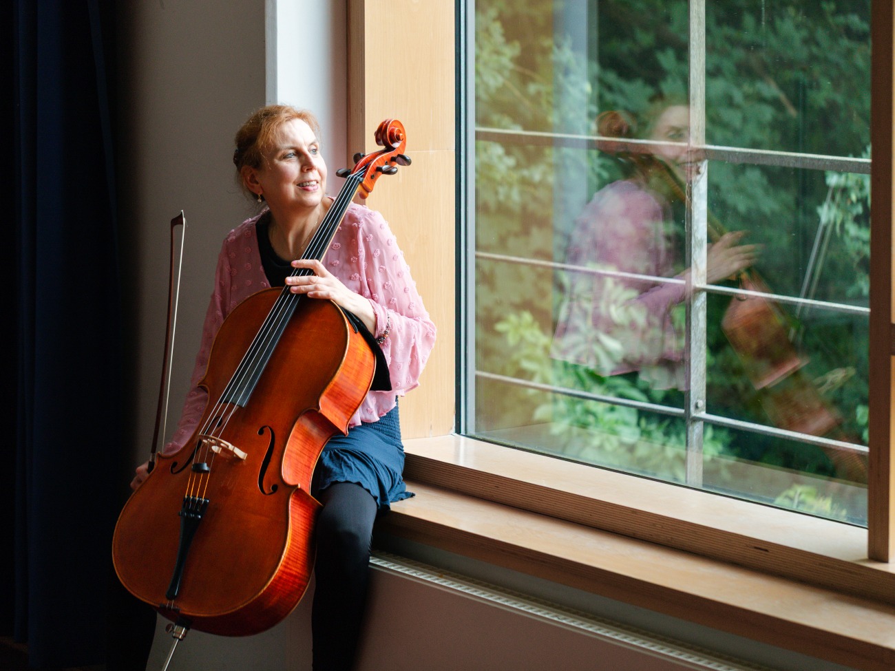 A woman is sitting on a windowsill with a cello and is looking out the window. 