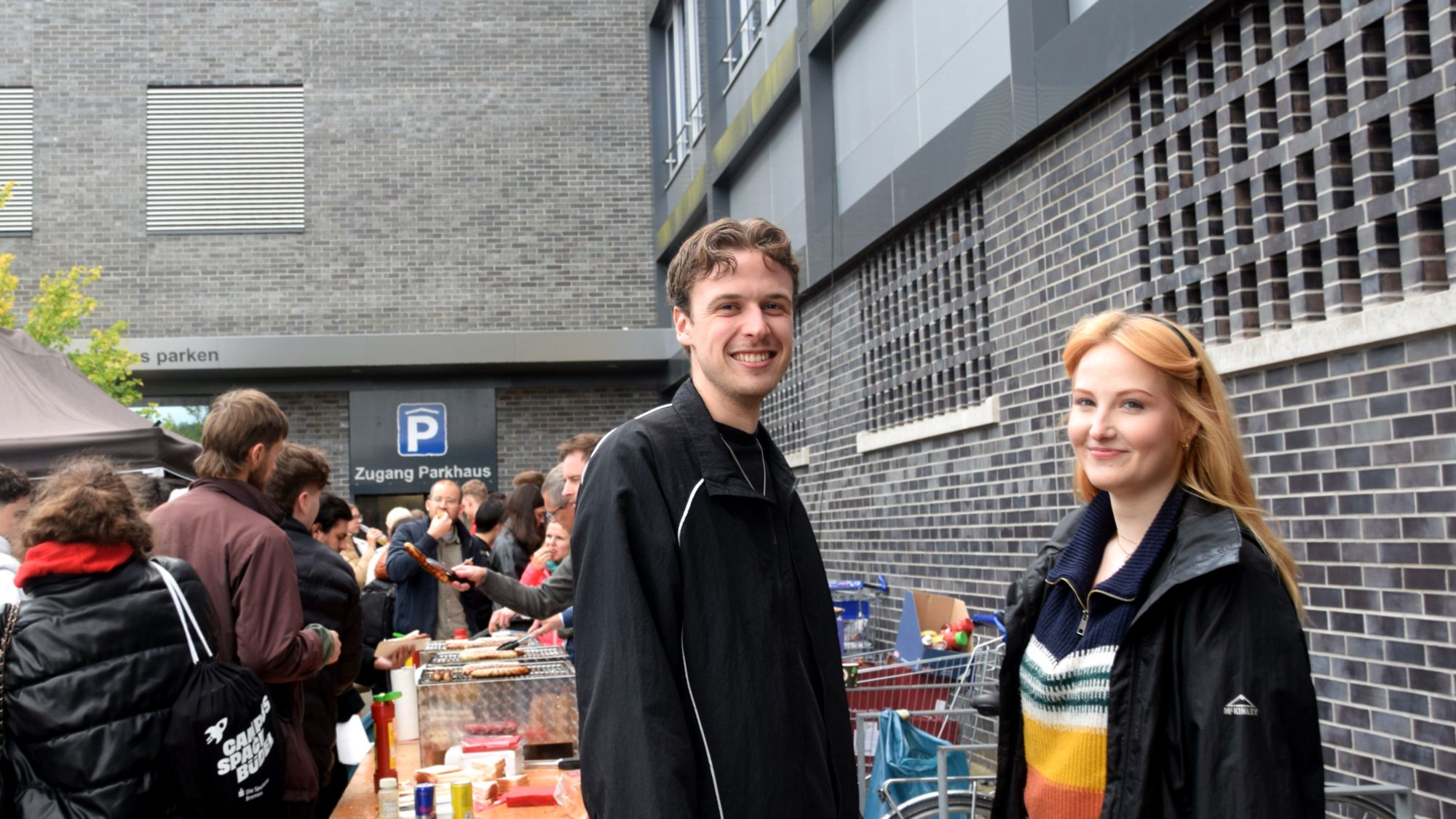 Zwei Personen lächeln in die Kamera. Im HIntergrund sieht man eine Schlange, die bei den Grills ansteht.