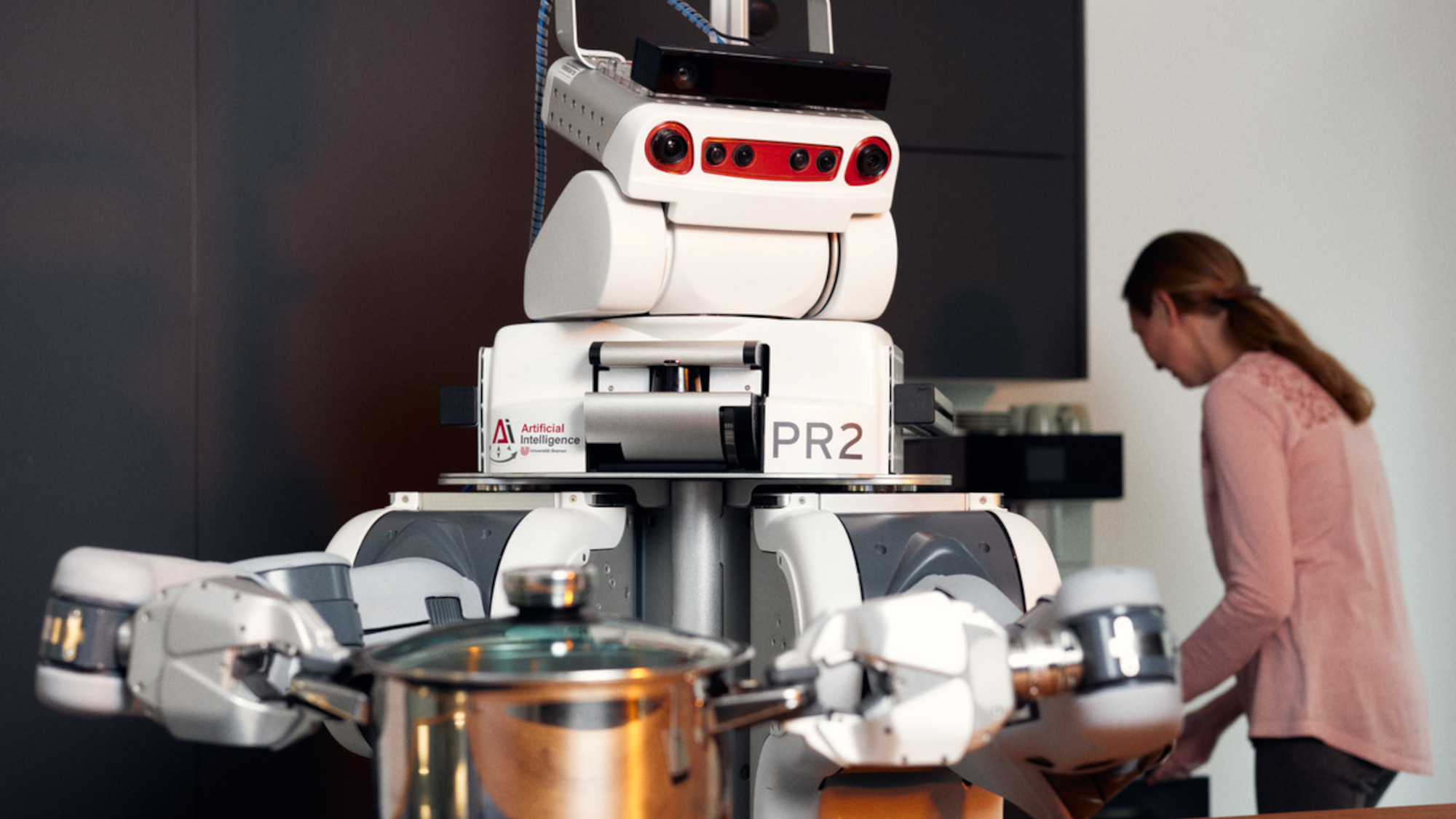 A robot holds a saucepan in its hands. It is helping the woman, who can be seen in the background, to cook.