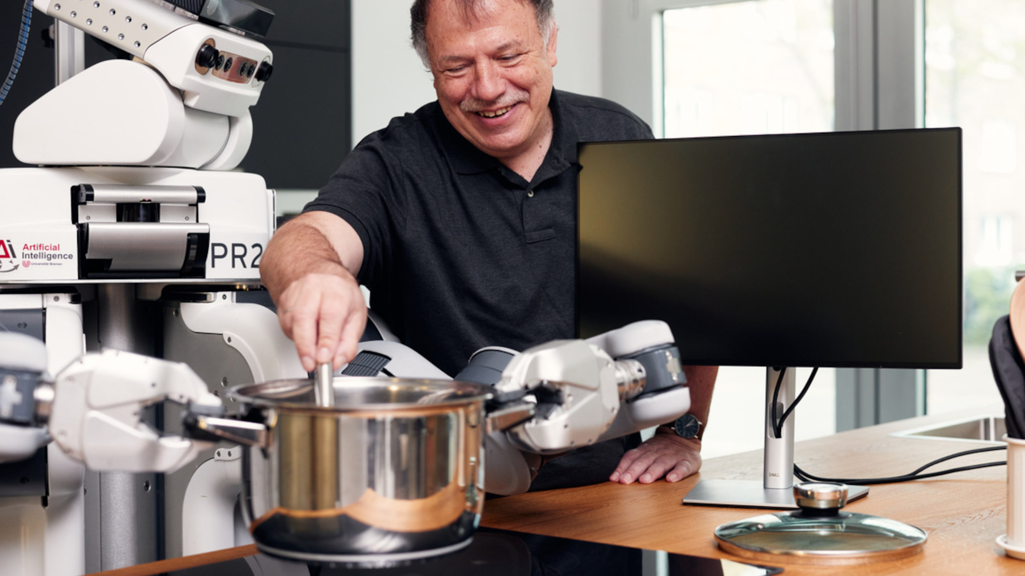 A robot holds a saucepan over the stove, Michael Beetz looks over its shoulder and stirs in the pot.