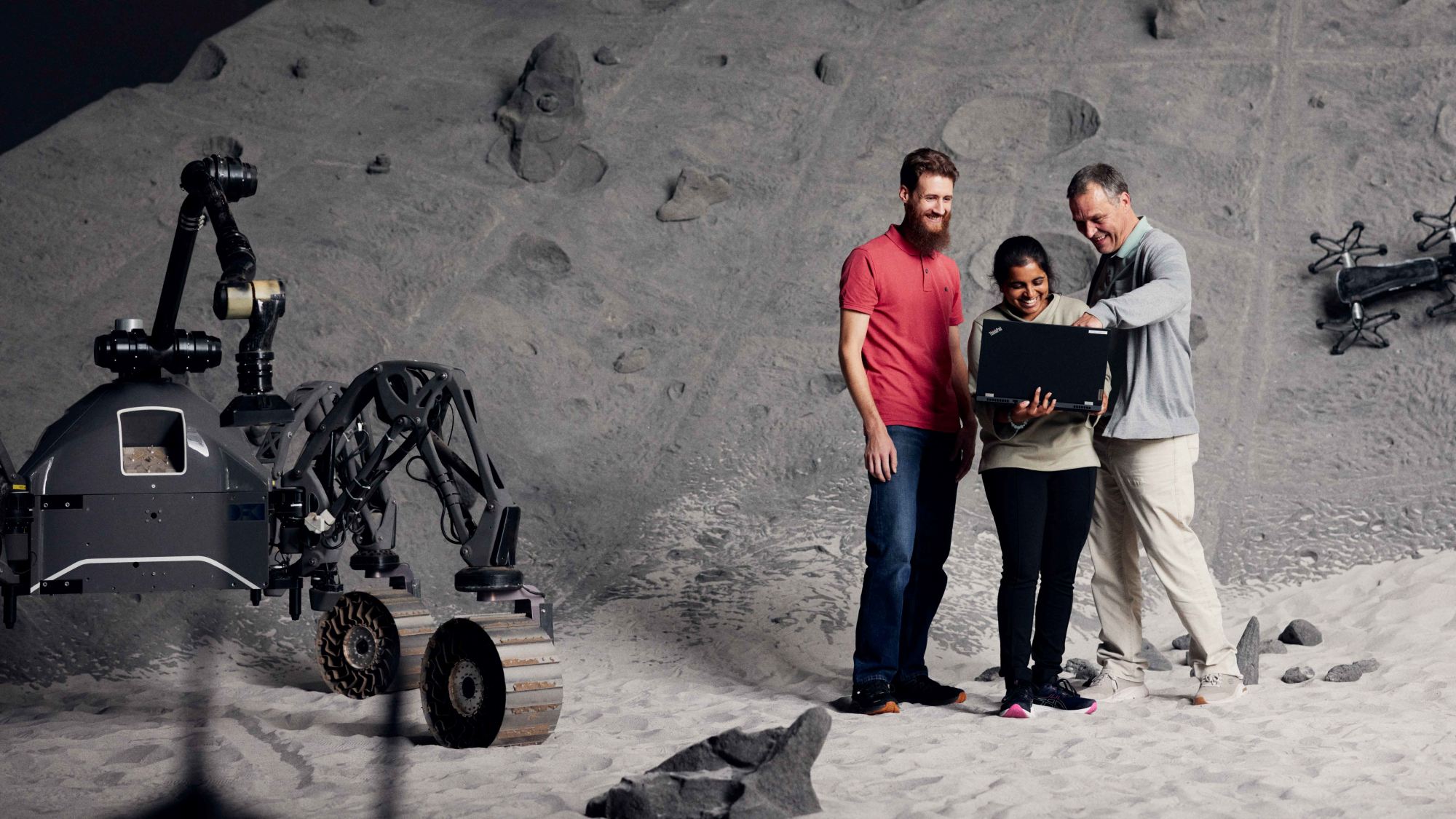 Three people stand next to a machine in a Mars-like landscape, looking at a laptop.