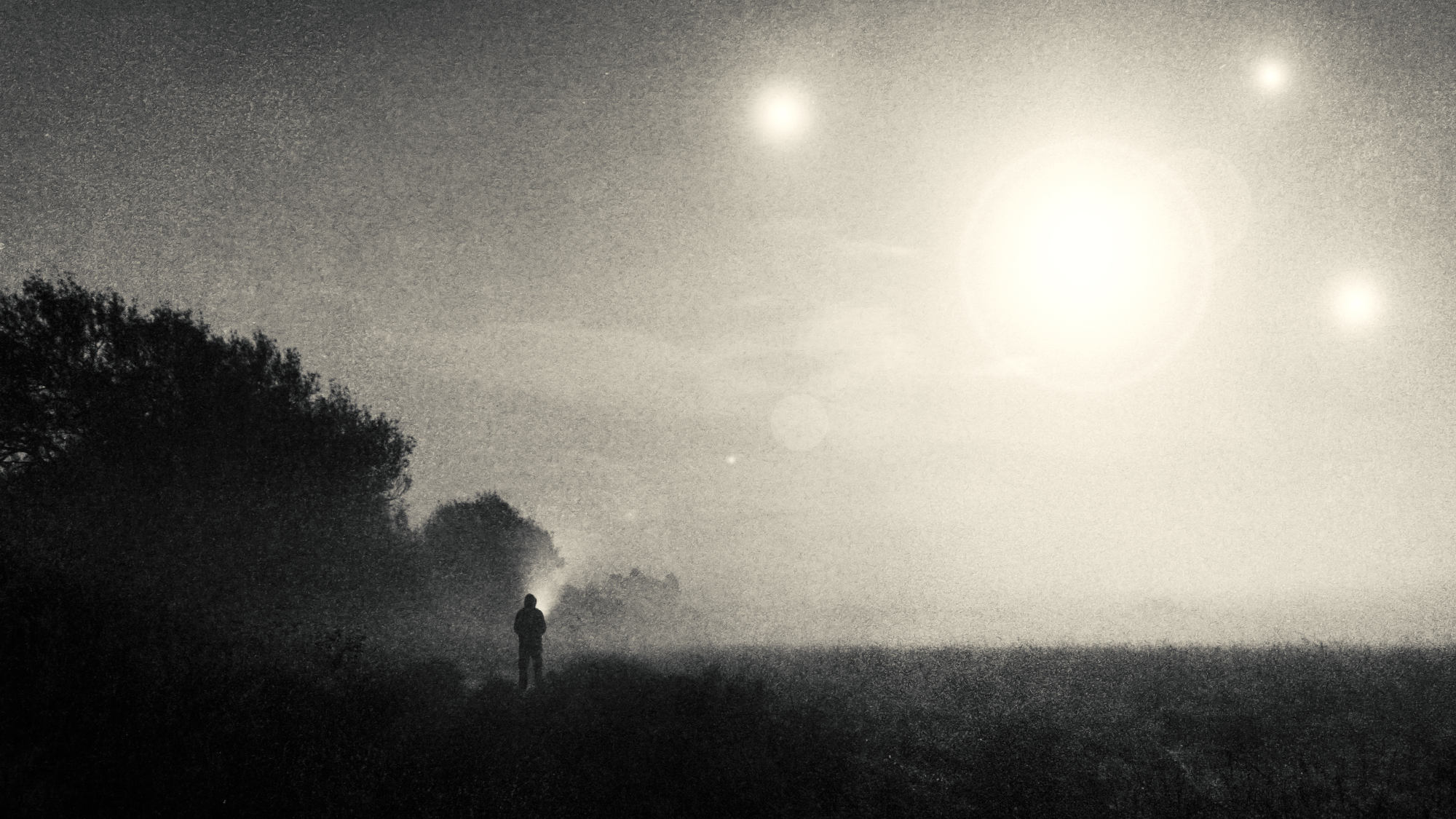 A person walks across a field with a flashlight; four differently sized points of light shine in the sky.