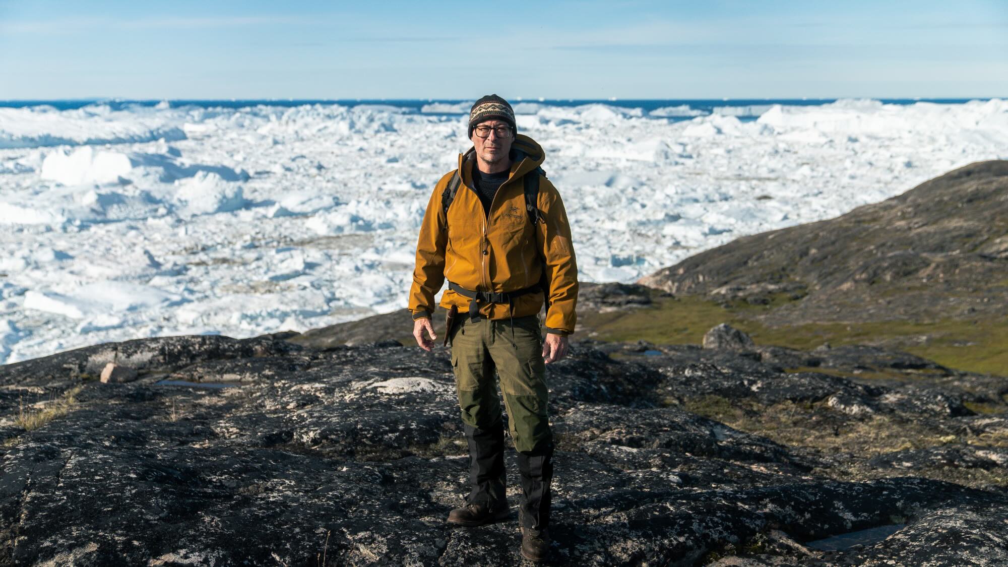 Professor Eske Willerslev is standing on a mountain, dressed in hiking gear. A landscape of ice and snow can be seen in the background.