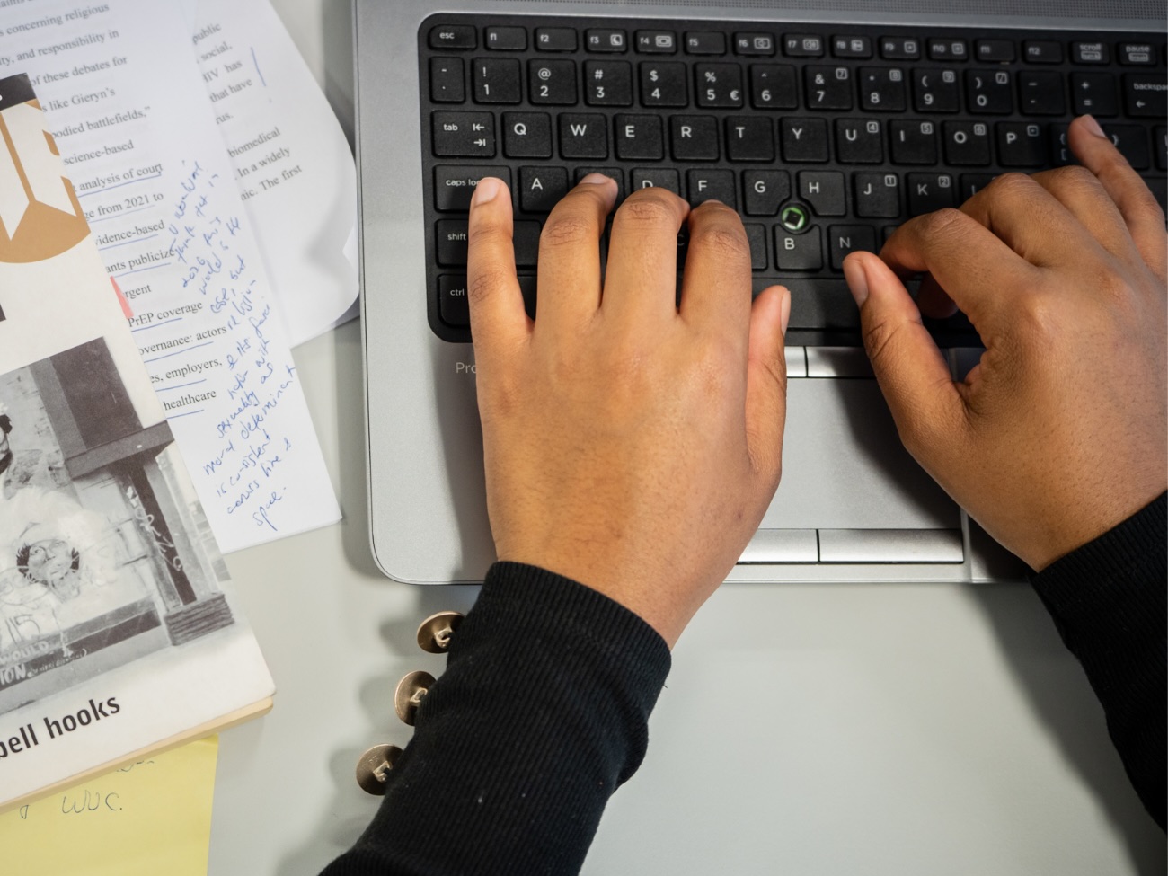 Das Bild zeigt einen Blick von oben auf den Ausschnitt eines Schreibtisches. Man sieht darin zwei Hände, die auf eine Laptop-Tastatur tippen. Neben der Tastatur liegt das Buch „Ain’t I a Woman“ von Bell Hooks auf einem kleinen Haufen von annotierten Texte