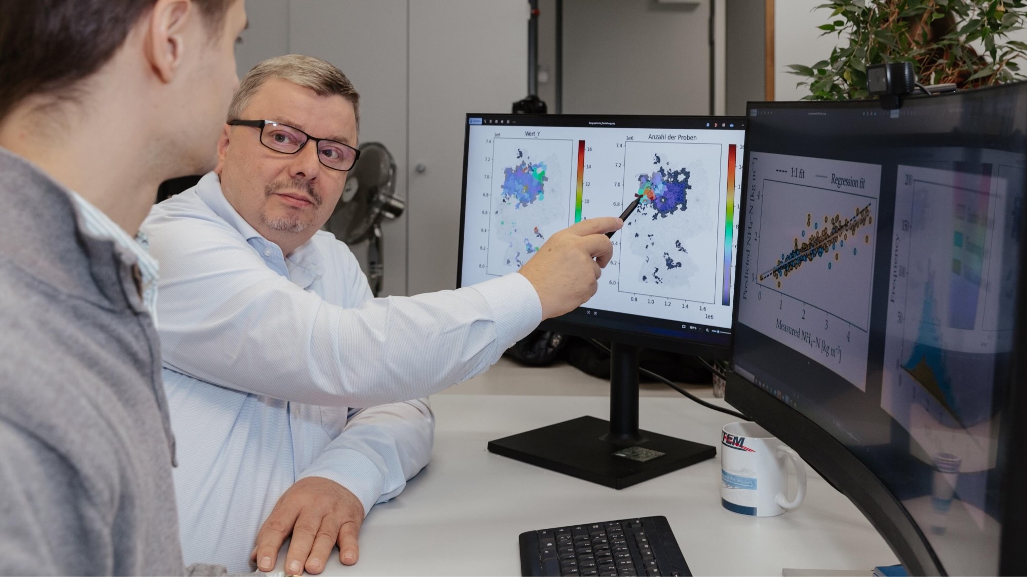 Eine Person zeigt einer anderen Person Messwerte auf einem der zwei ihnen gegenüberstehenden Bildschirme.