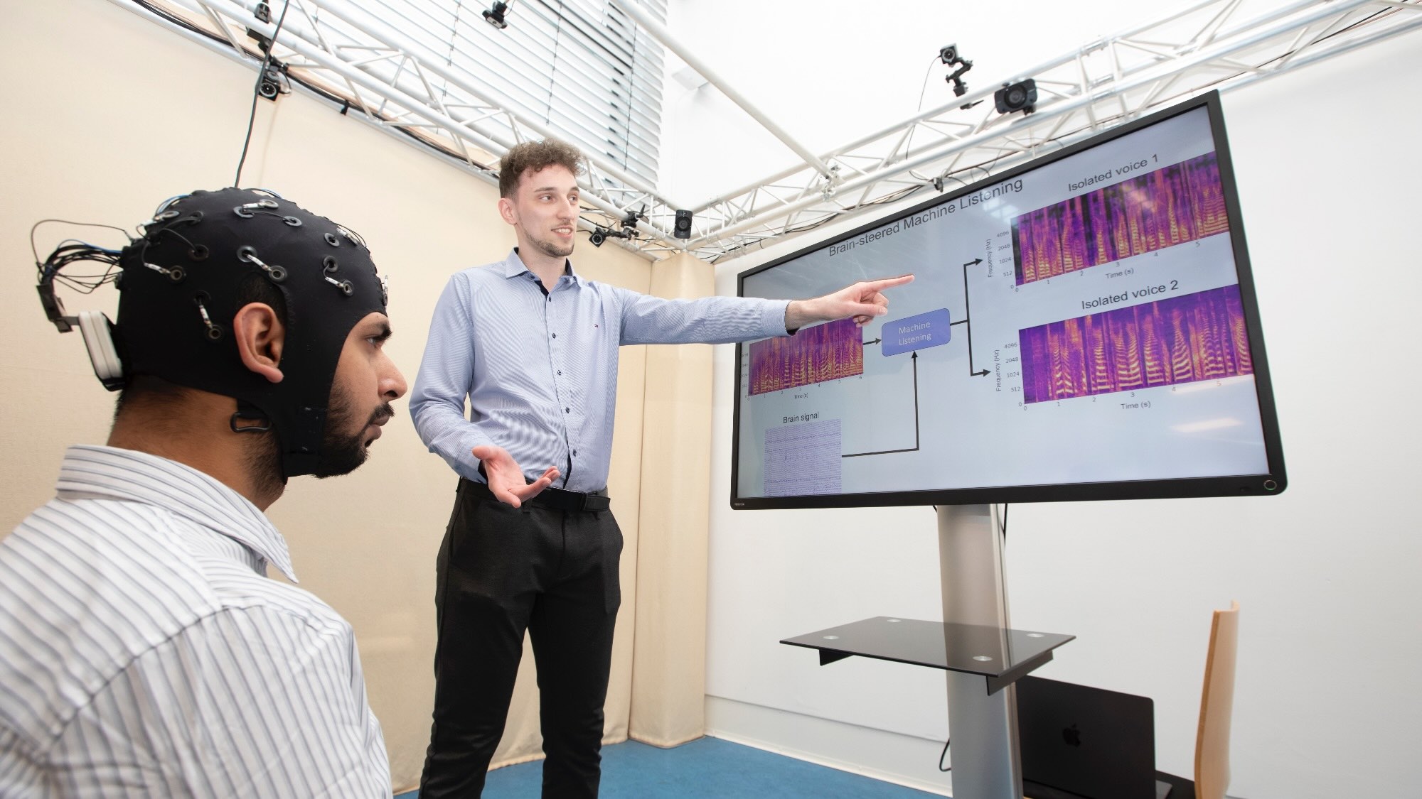 The picture shows two people. One of them is pointing at an infographic on a screen with the English headline “Brain-steered Machine Listening.” Seated in front of that person is another man looking at the screen. He is wearing a hood equipped with several cables.
