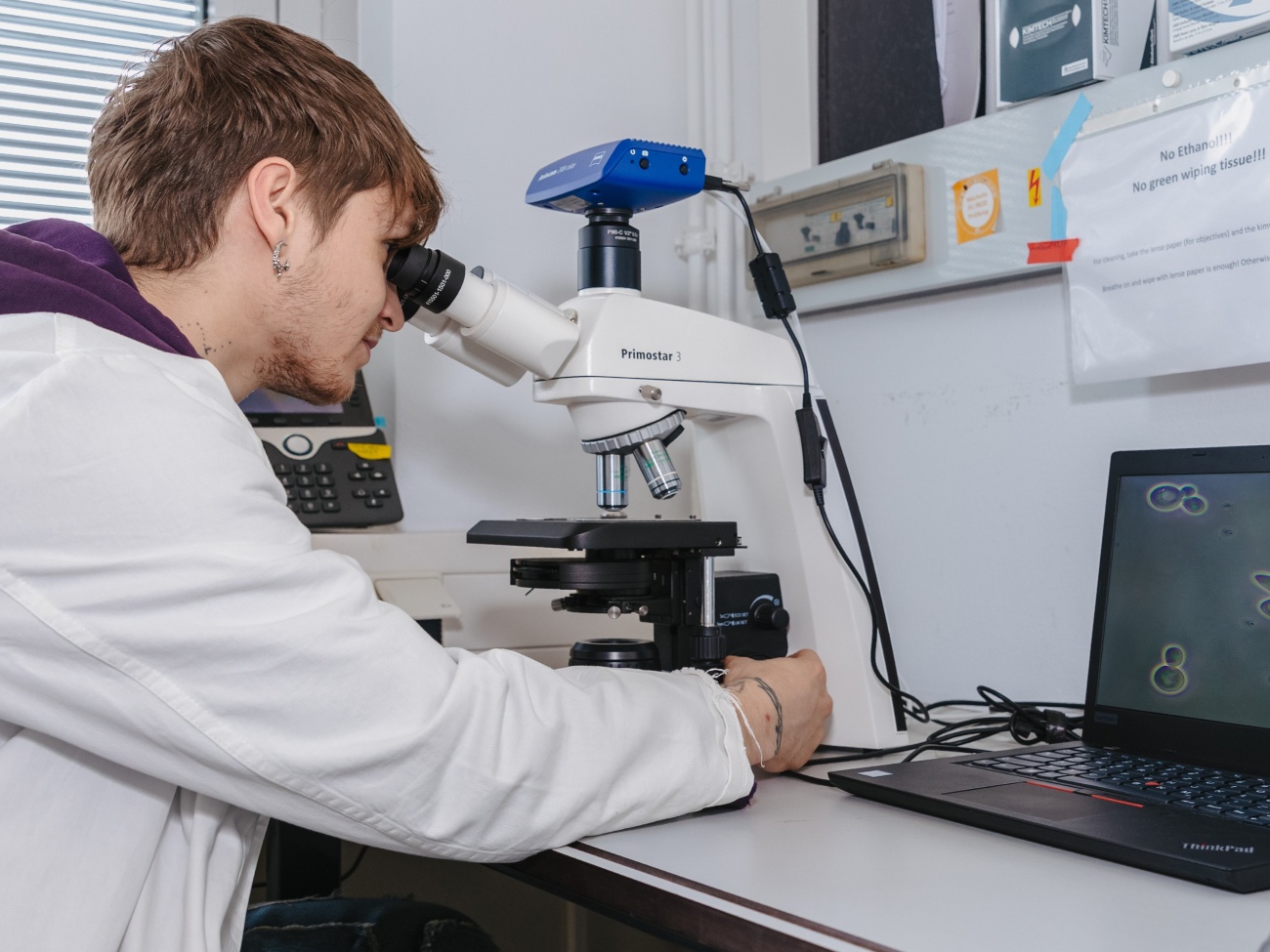 A person wearing a lab coat looks through a microscope.