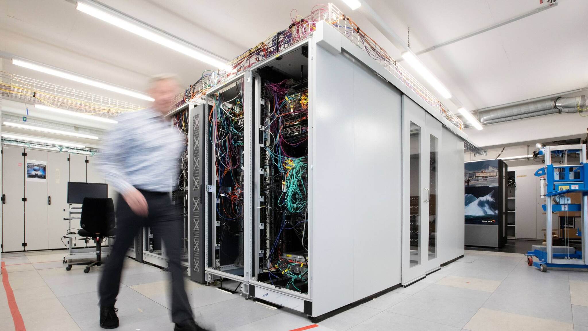 The picture shows a server room in which a person walks past tall computer equipment filled with colorful cables.