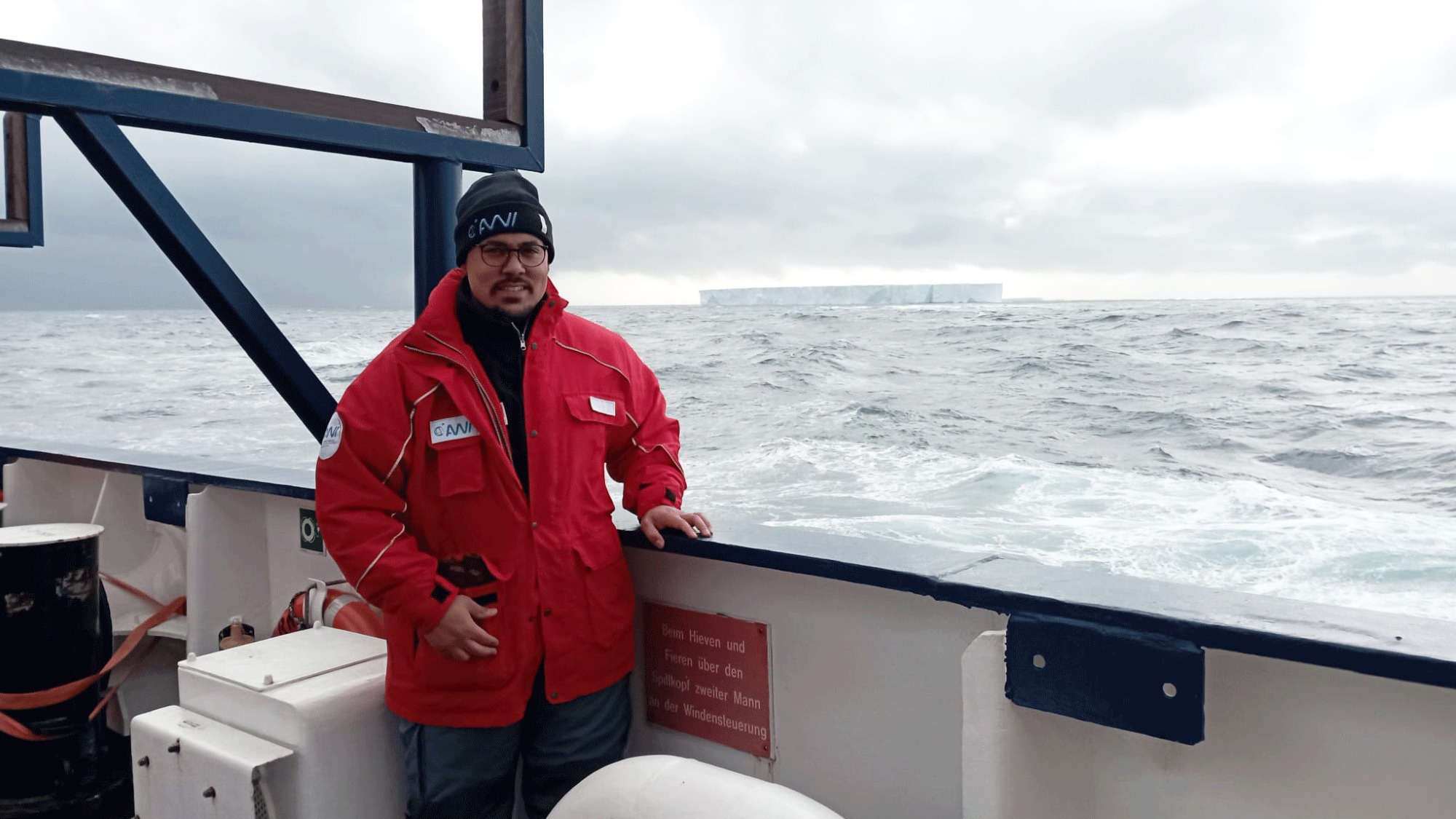 In the foreground, you can see someone standing on the deck of a boat wearing a thick winter jacket. In the background, you can see an iceberg rising above the water.