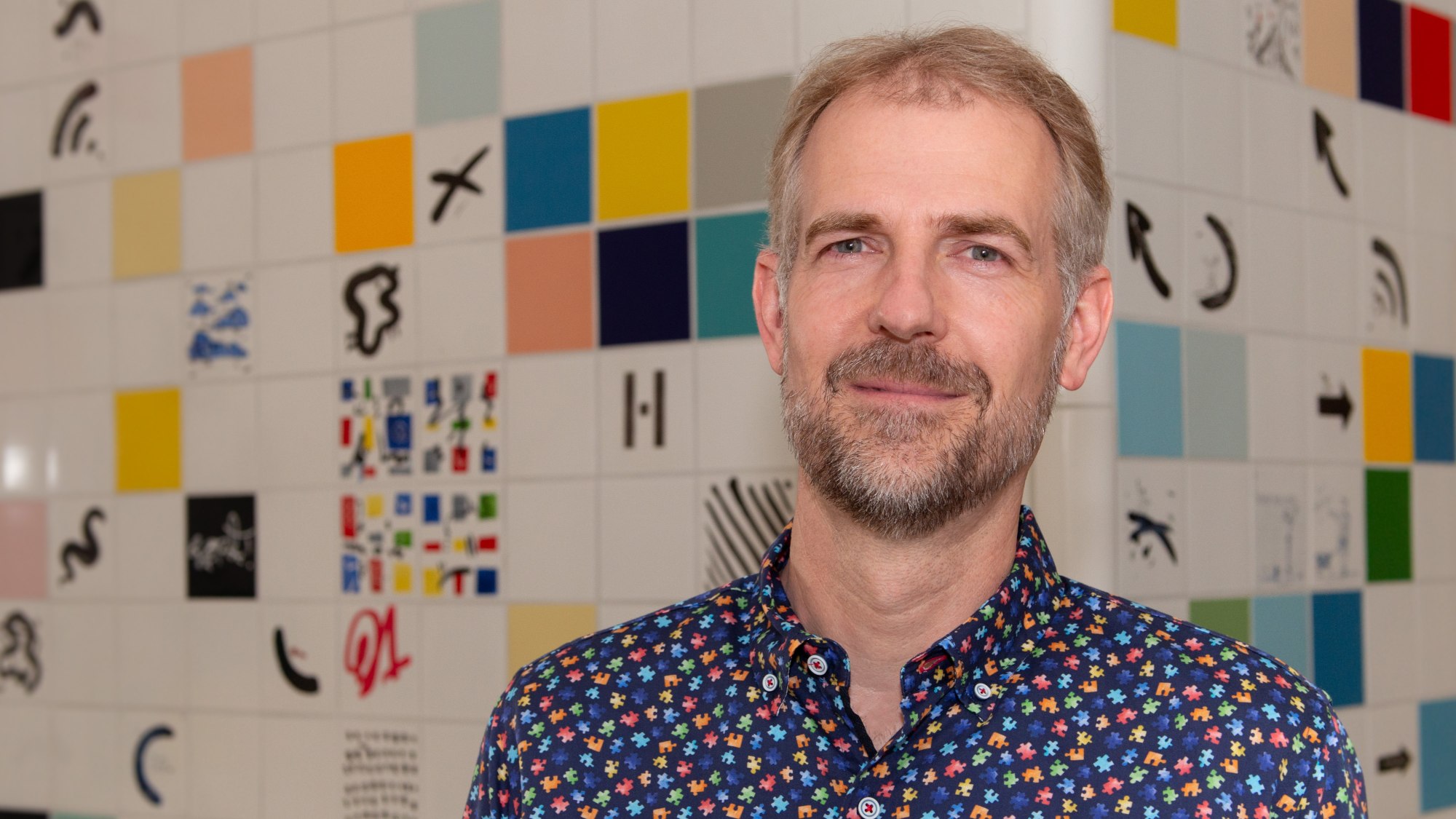 Professor Norman Sieroka stands in front of a colorful tiled wall wearing a shirt decorated with puzzle pieces.