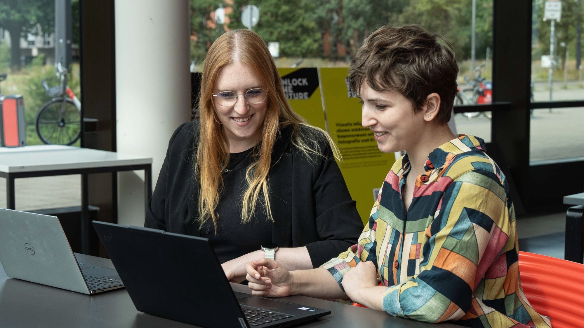 Caroline Dominik and Nicola Thoben are sitting together in front of two laptops and looking at one of the two screens.