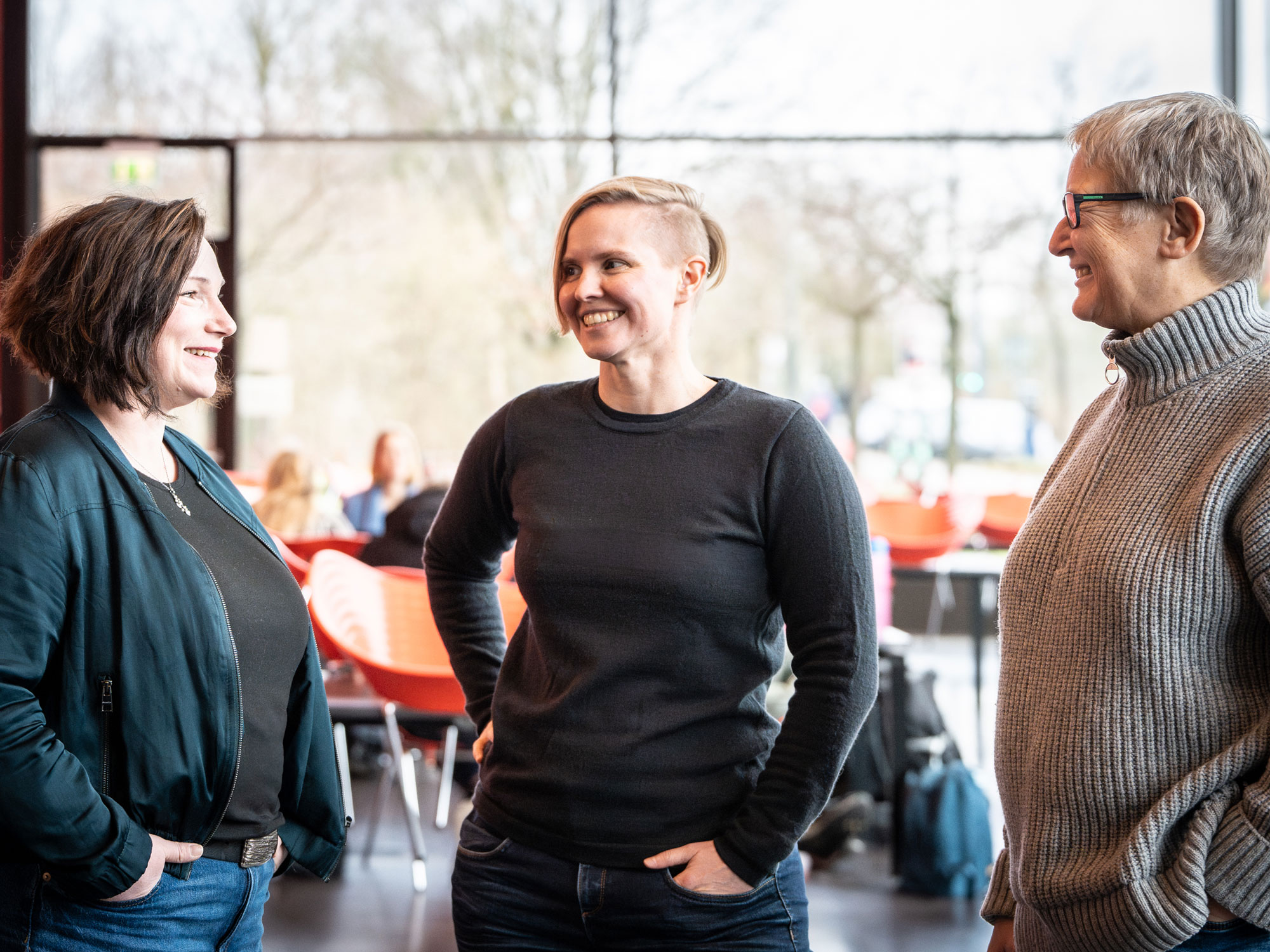 Three people are standing in a modern, well-lit room, smiling and engaged in conversation. They appear relaxed and involved. In the background, more people are sitting at tables, creating a casual and collaborative atmosphere.