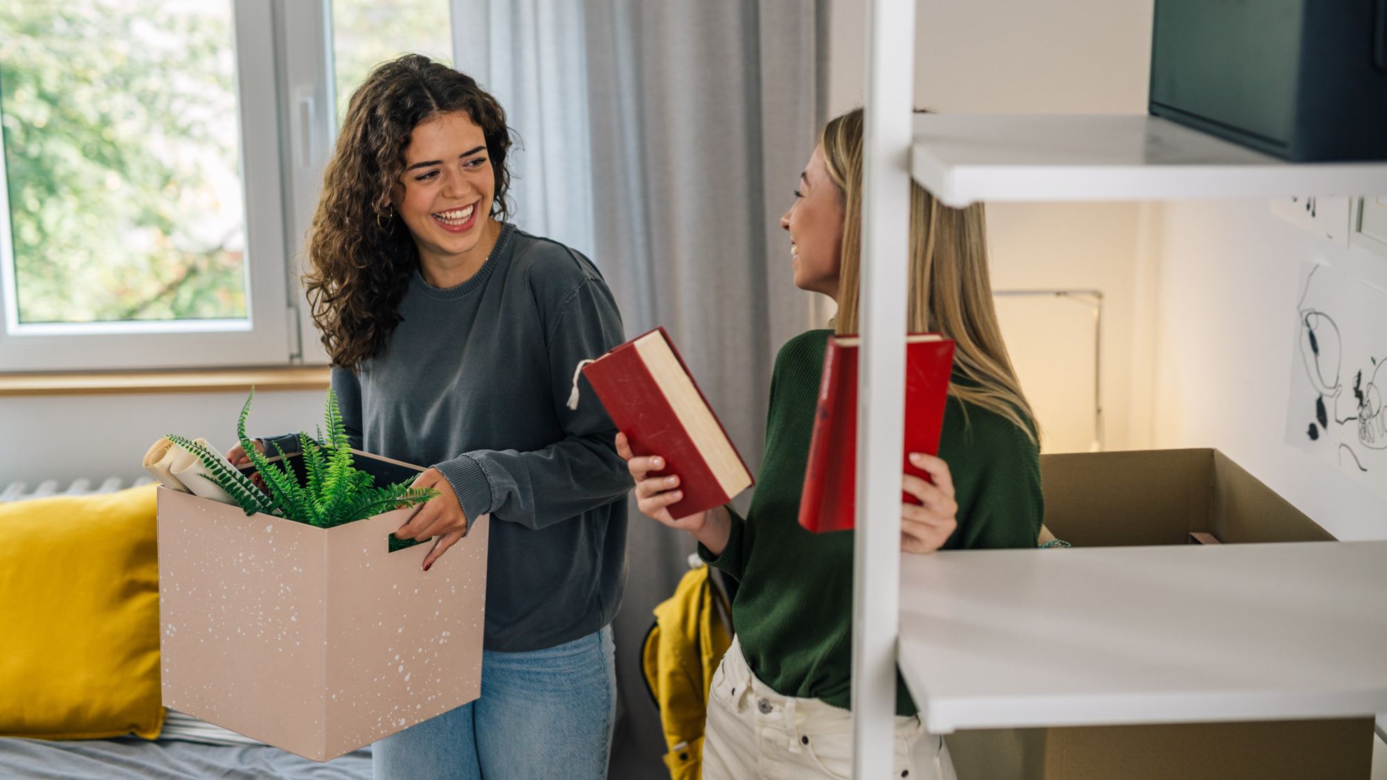 A woman is standing in a room with a moving box and is smiling at another woman holding two books.