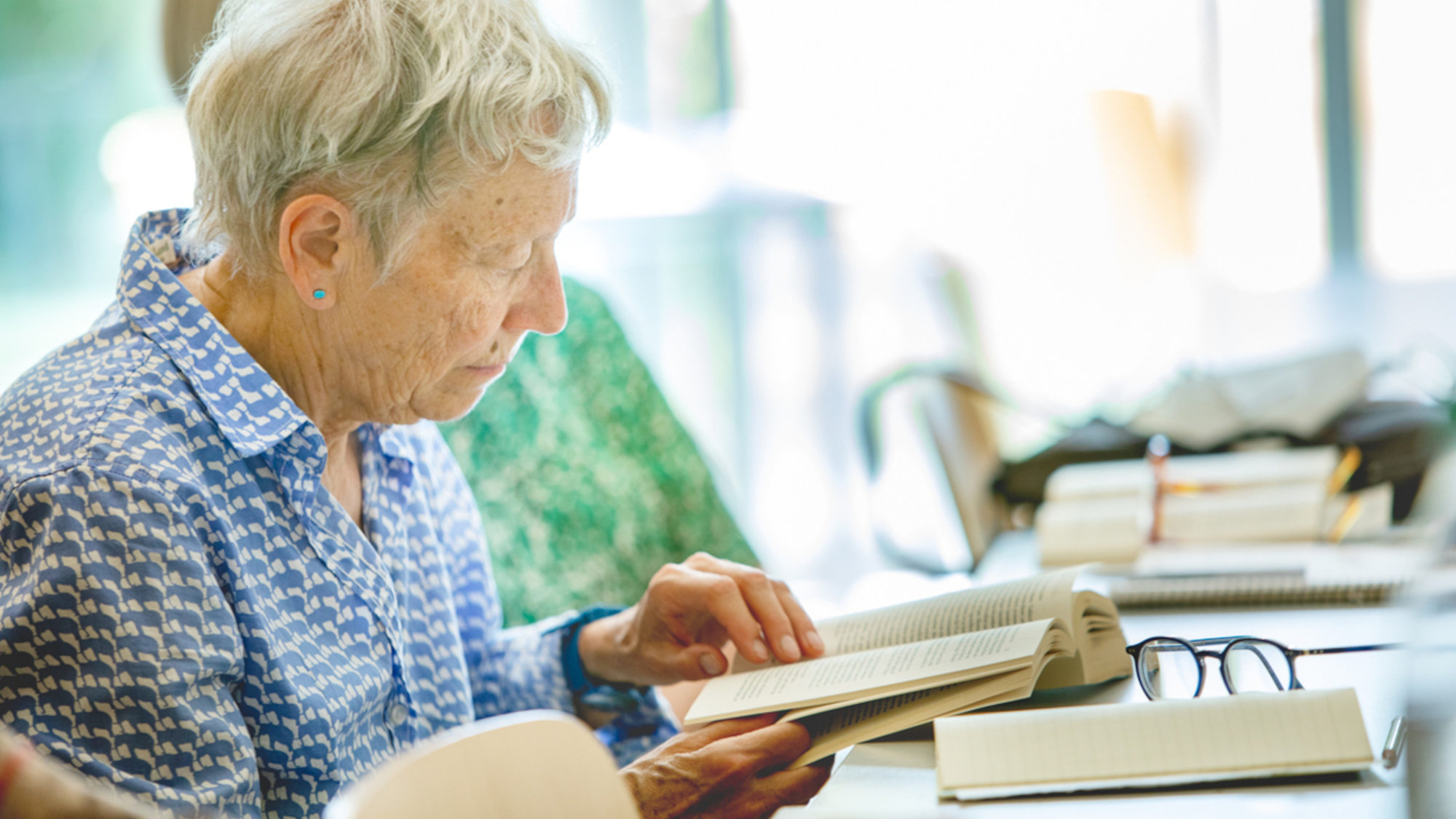 A senior student is reading in a book, with other books next to her.