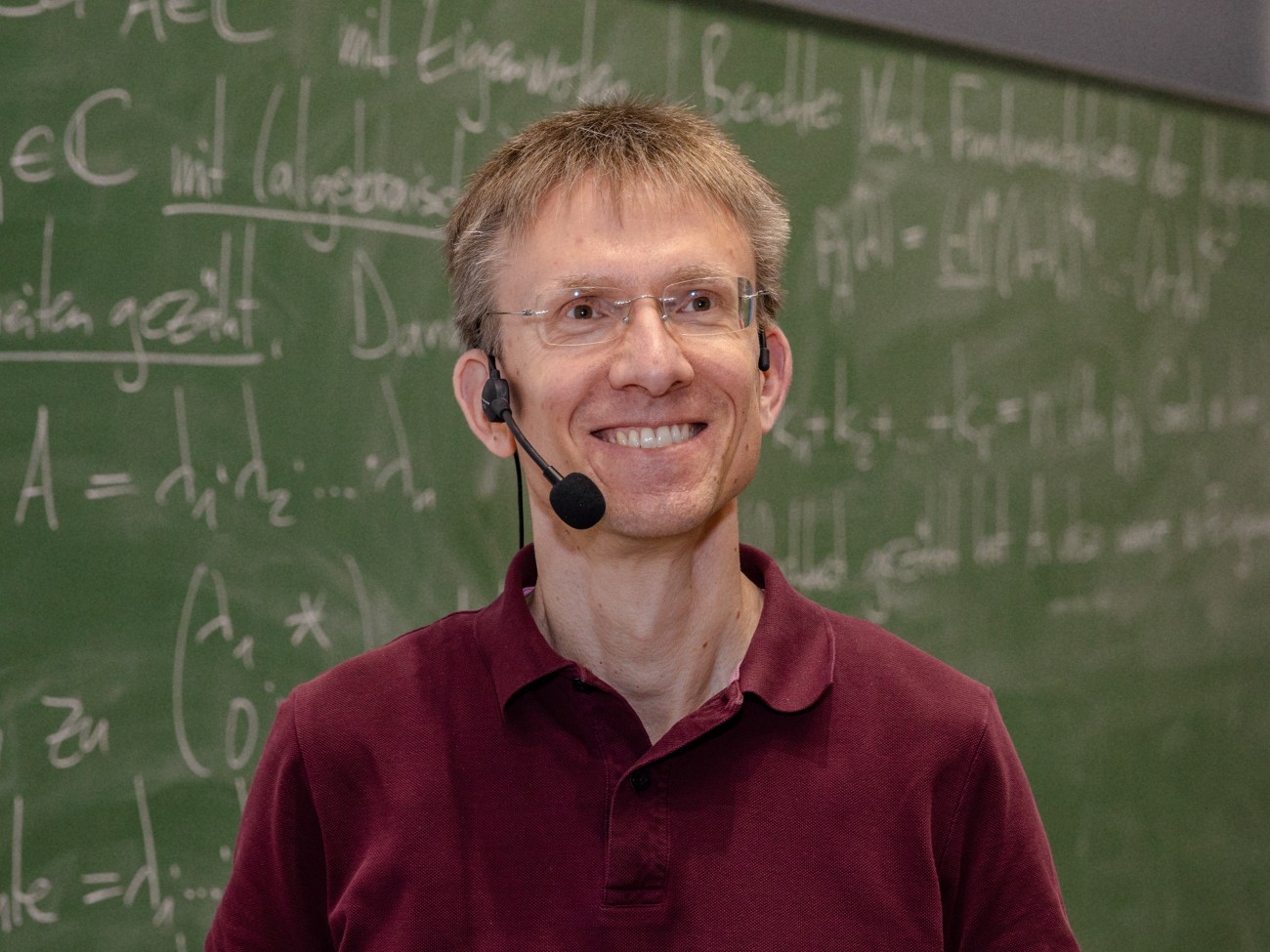 Ein Professor mit einem Headset lächelt vor einer Tafel in die Kamera.