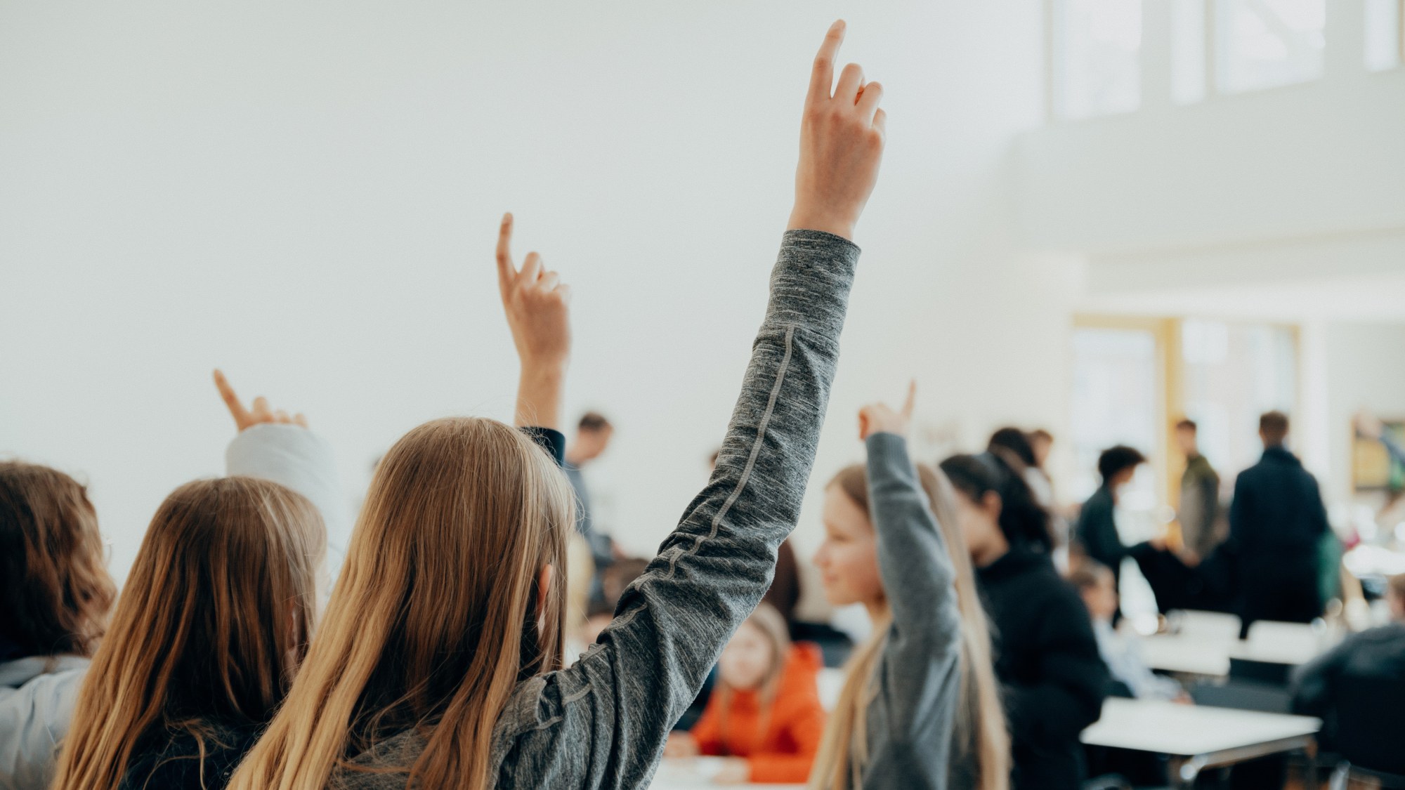 Several children are sitting at tables in a room and raising their hands.