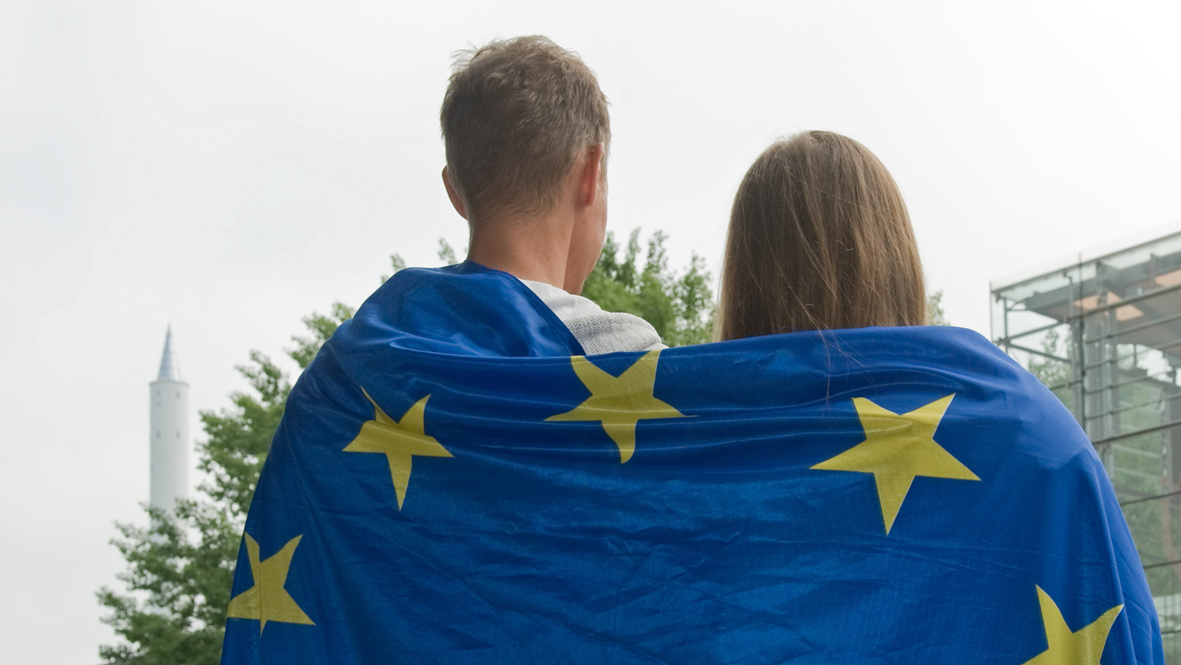 Zwei Personen von hinten haben eine Europaflagge um die Schultern gelegt und stehen vor der Glashalle der Universität Bremen.