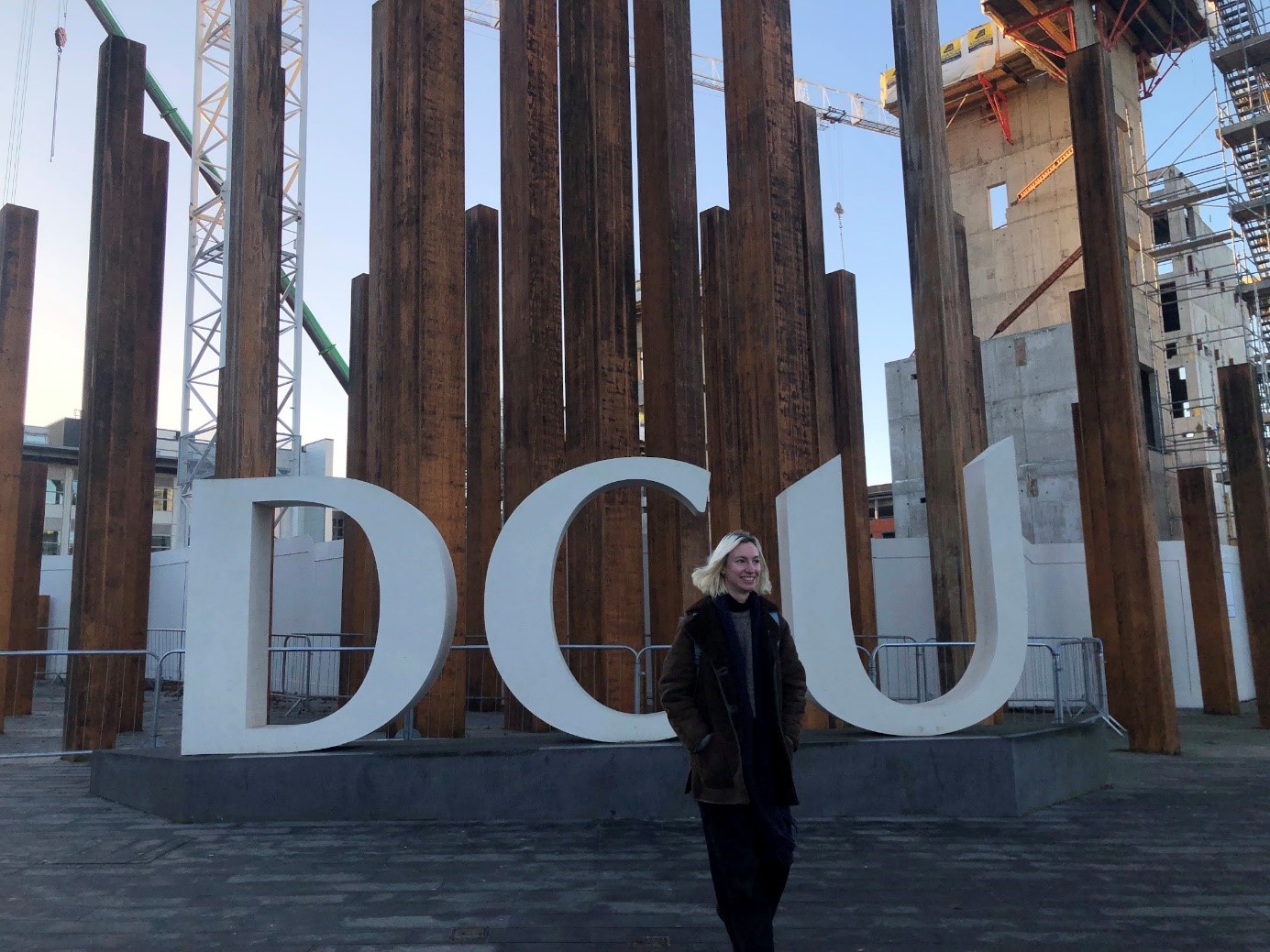Eine junge Studentin steht vor dem rostroten Säulen und dem Schriftzug DCU.
