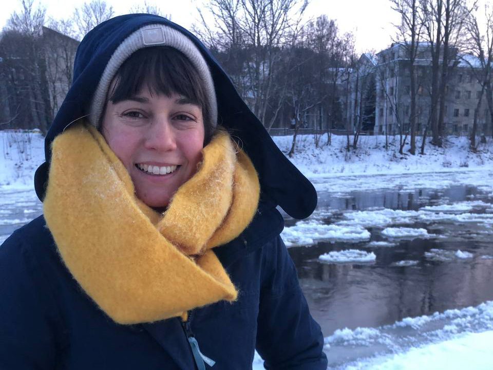 Eine junge Studentin mit gelben Schal steht in einer Winterlandschaft vor einem Fluss.