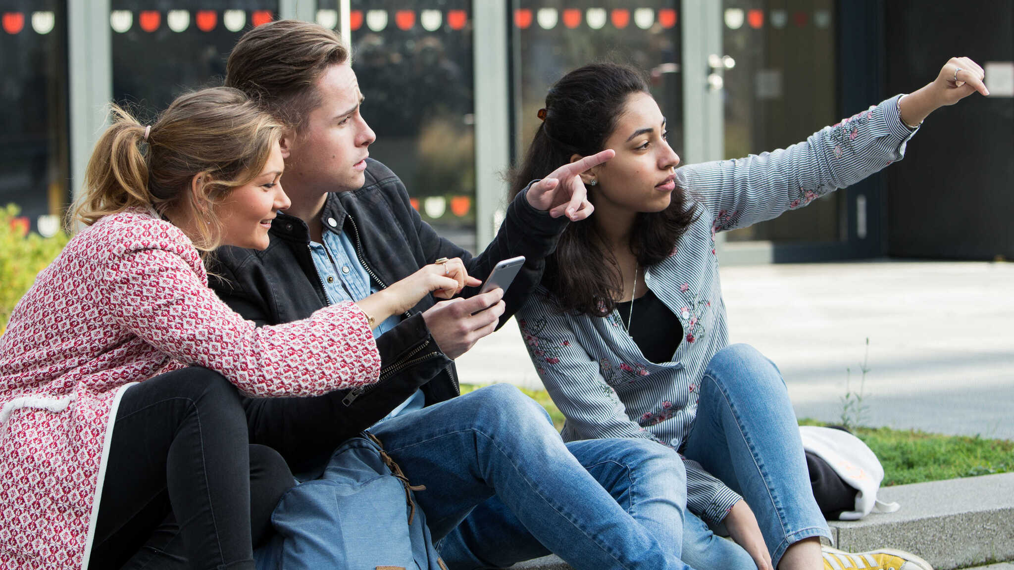 Drei Studierende sitzen draußen auf dem Campus. Sie schauen in die Richtung, in die zwei von ihnen zeigen.