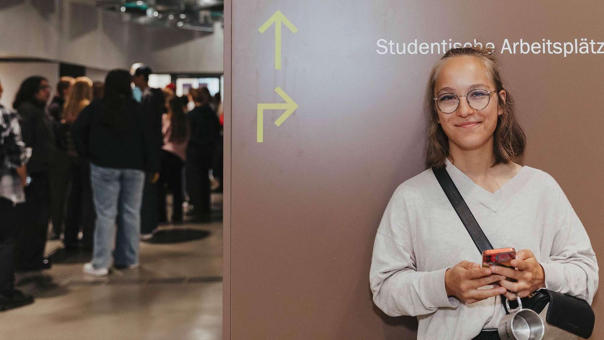 Eine junge Person steht mit einem Smartphone in der Hand vor einer Wand versehen mit der Aufschrift „Studentische Arbeitsplätze“ und zwei Richtungspfeilen und lächelt in die Kamera. Neben der Wand ist eine Schlange von Menschen zu erkennen.