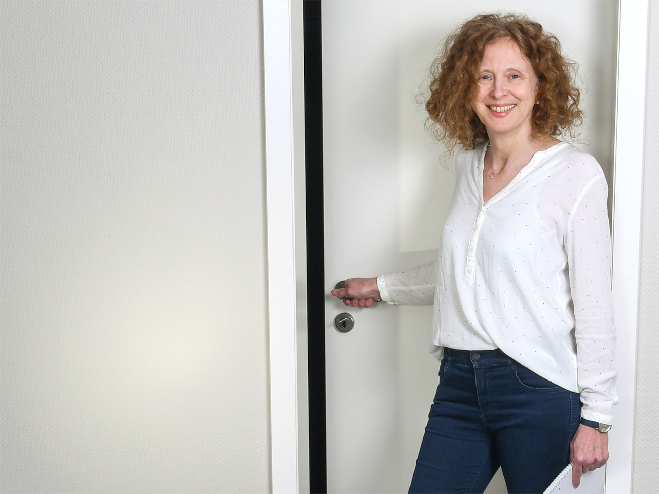 Die habilitierte Universitätslektorin Iris Stahlke steht vor einer weißen Tür. Sie hält die Türklinke in der Hand und schaut lächelnd in die Kamera.