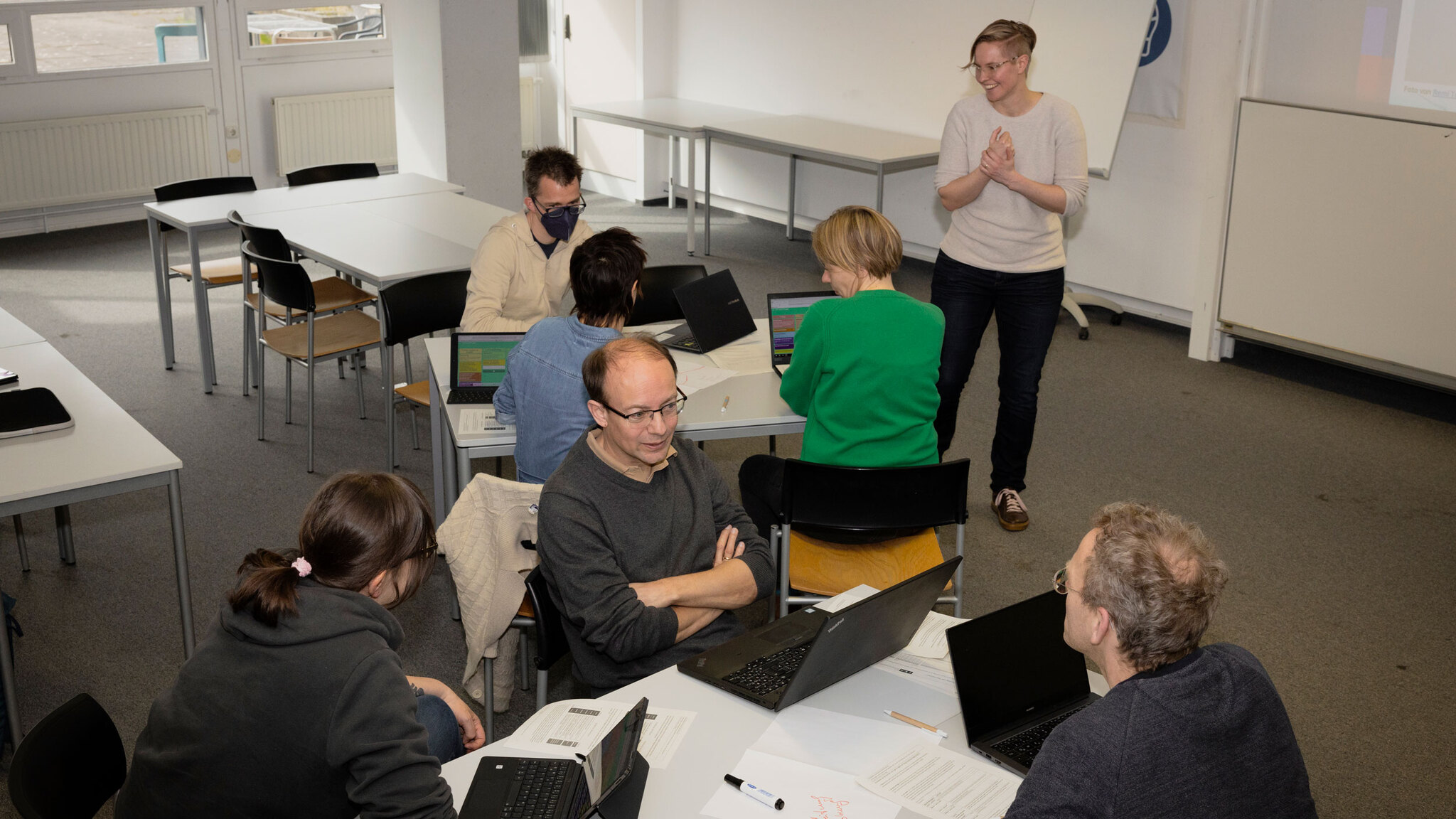 A group workspace with several people working on laptops and engaging in discussions. One person stands at the front, smiling and appearing to lead a presentation or discussion. The atmosphere seems cooperative and engaged. The room is equipped with tables, chairs, and whiteboards.