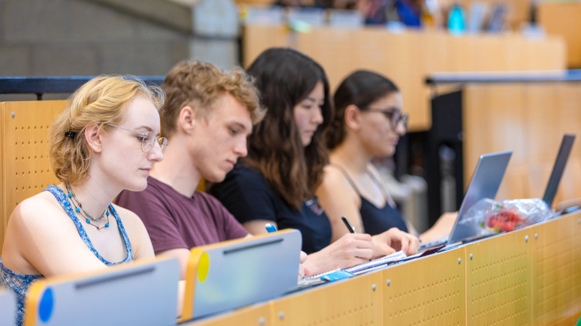 Vier Studierende sitzen im Hörsaal und arbeiten an ihren Laptops während einer Vorlesung