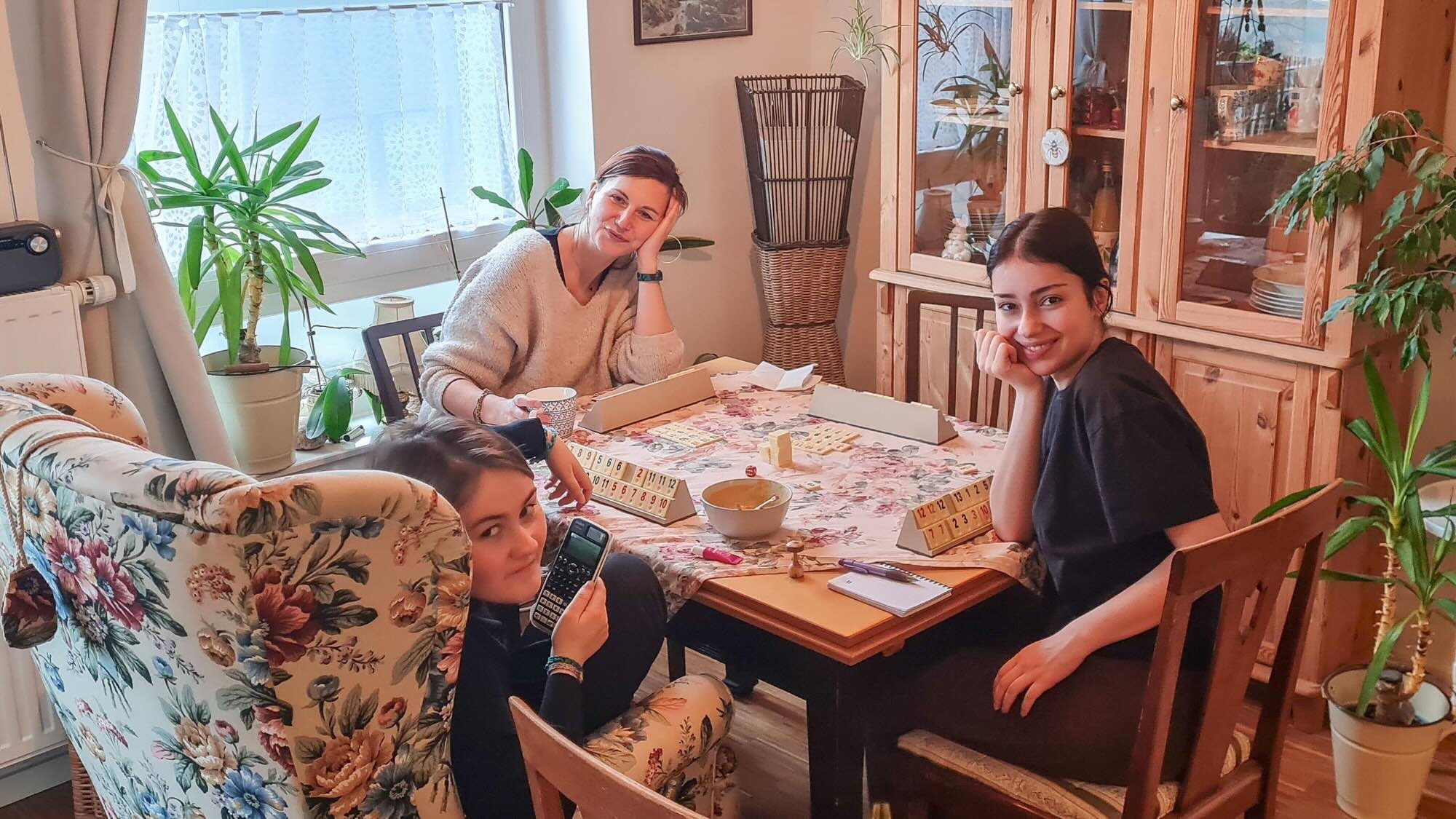 A picture of Zeynep, Britta Plote, and their daughter sitting around a table playing a game and smiling at the camera. 