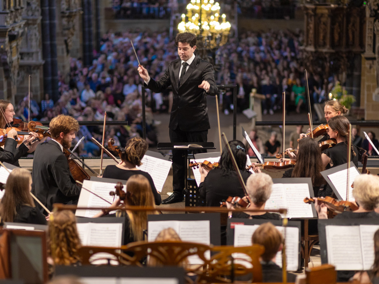 A male conductor stands in front of an orchestra.