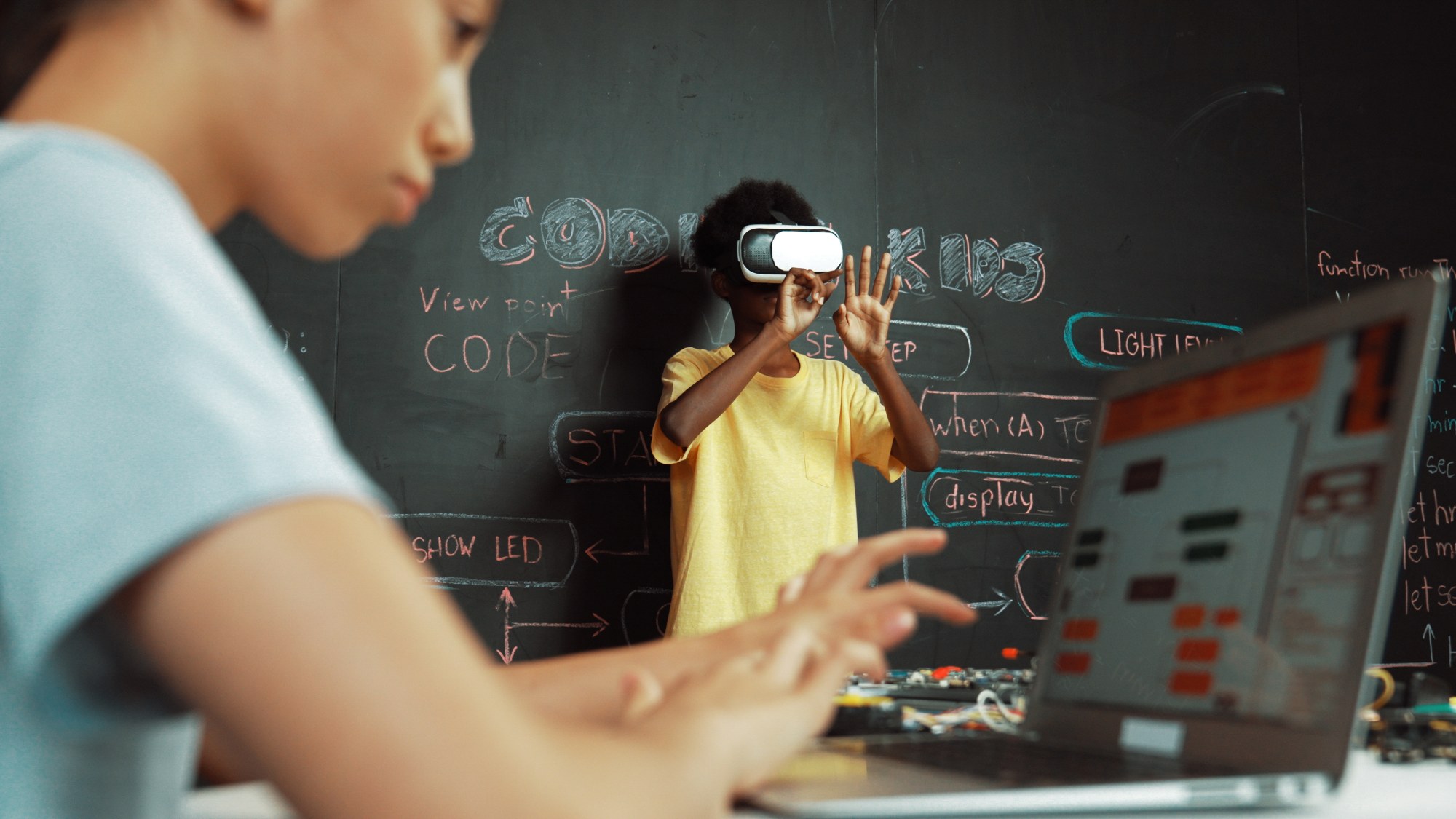 In the foreground, a child is sitting at a computer. In the background, another child is wearing VR glasses.