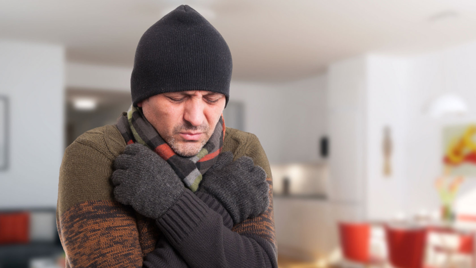 A man in a scarf and hat stands freezing in the living room, hugging himself.