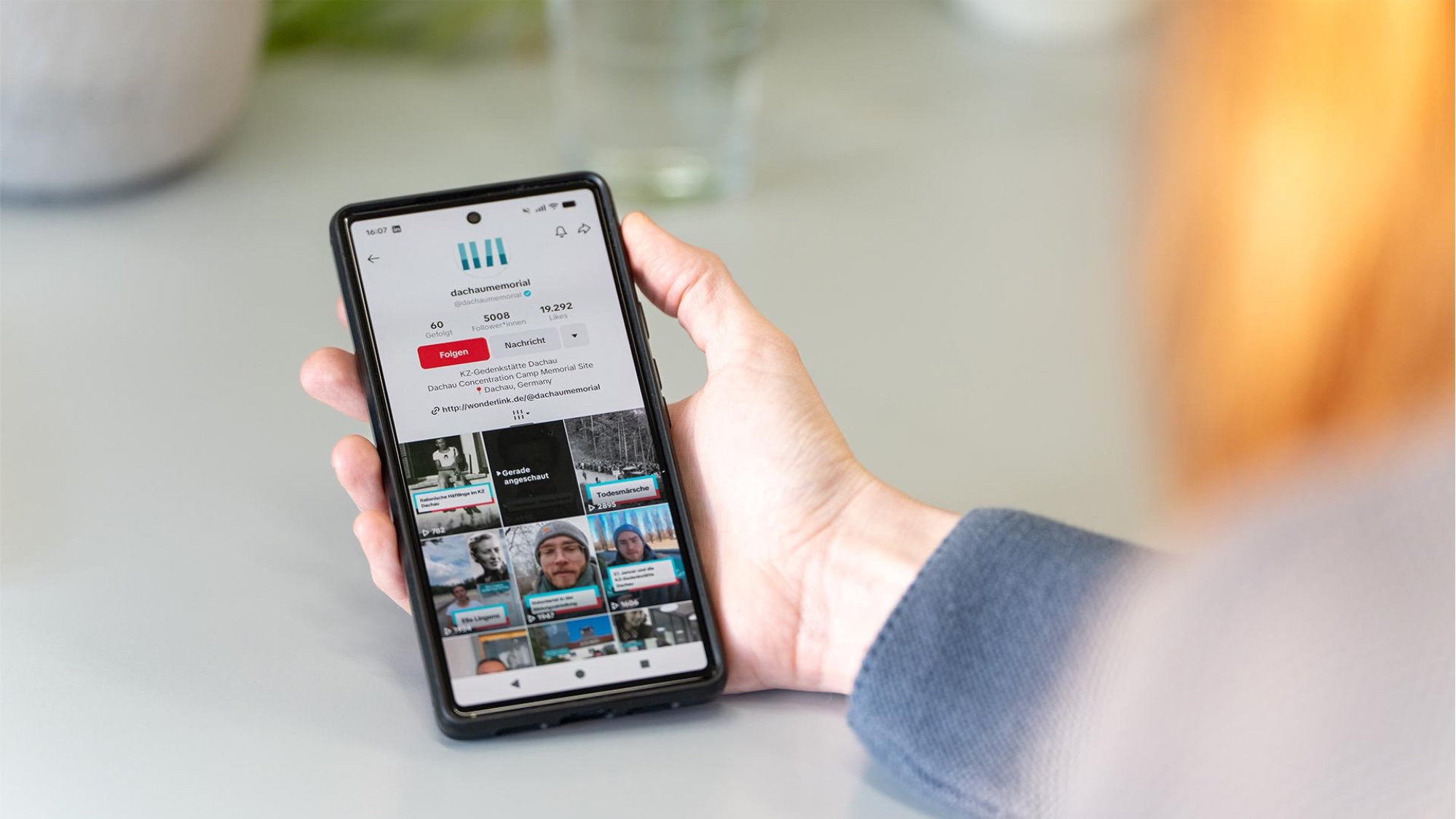The hand of a person's holding a phone. The phone screen displays the TikTok account of “dachaumemorial”.