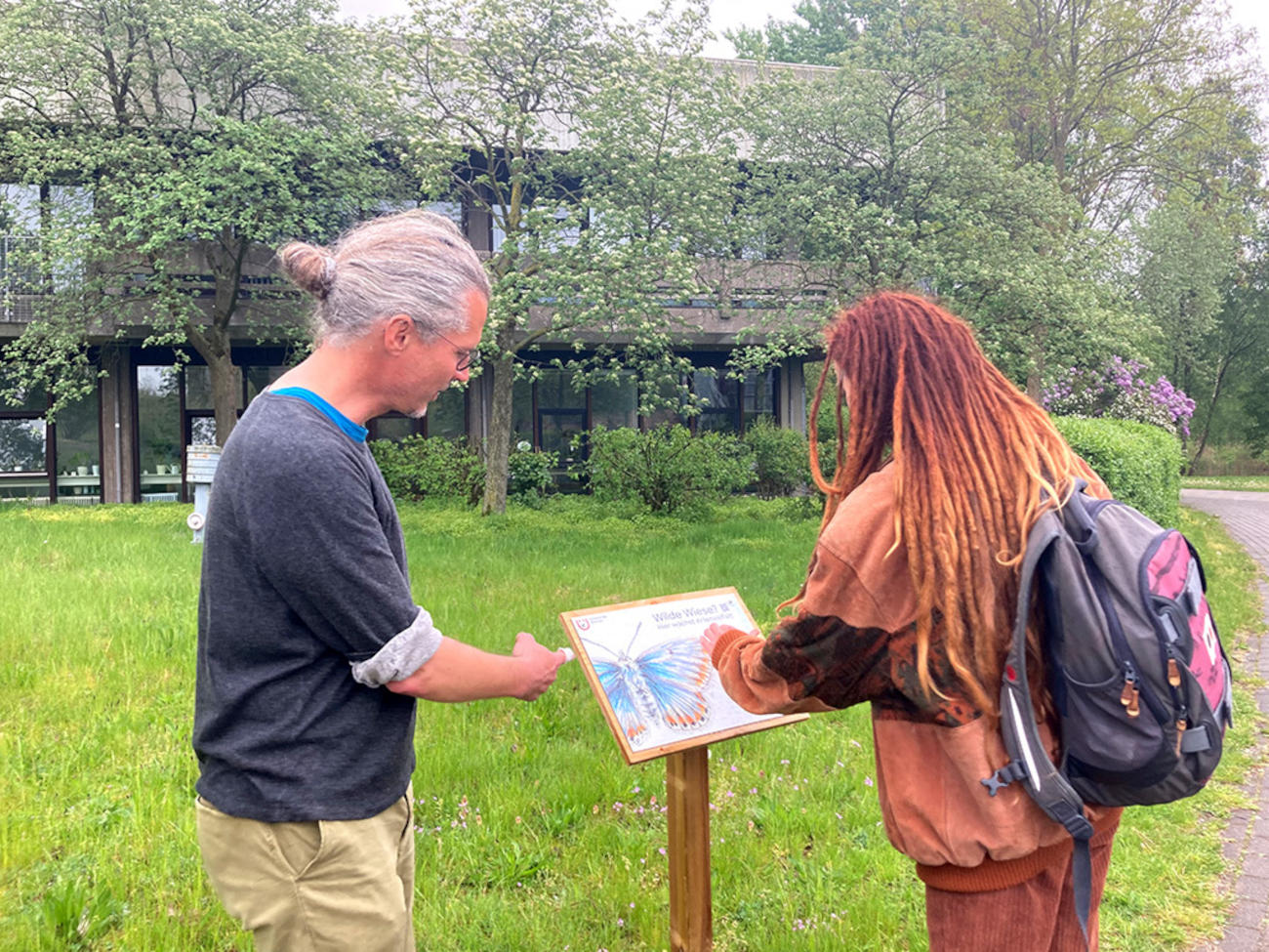 Marko Rohlfs and Lorena Kalvelage stand in front of one of their information boards next to a meadow where a large blue butterfly can be seen.