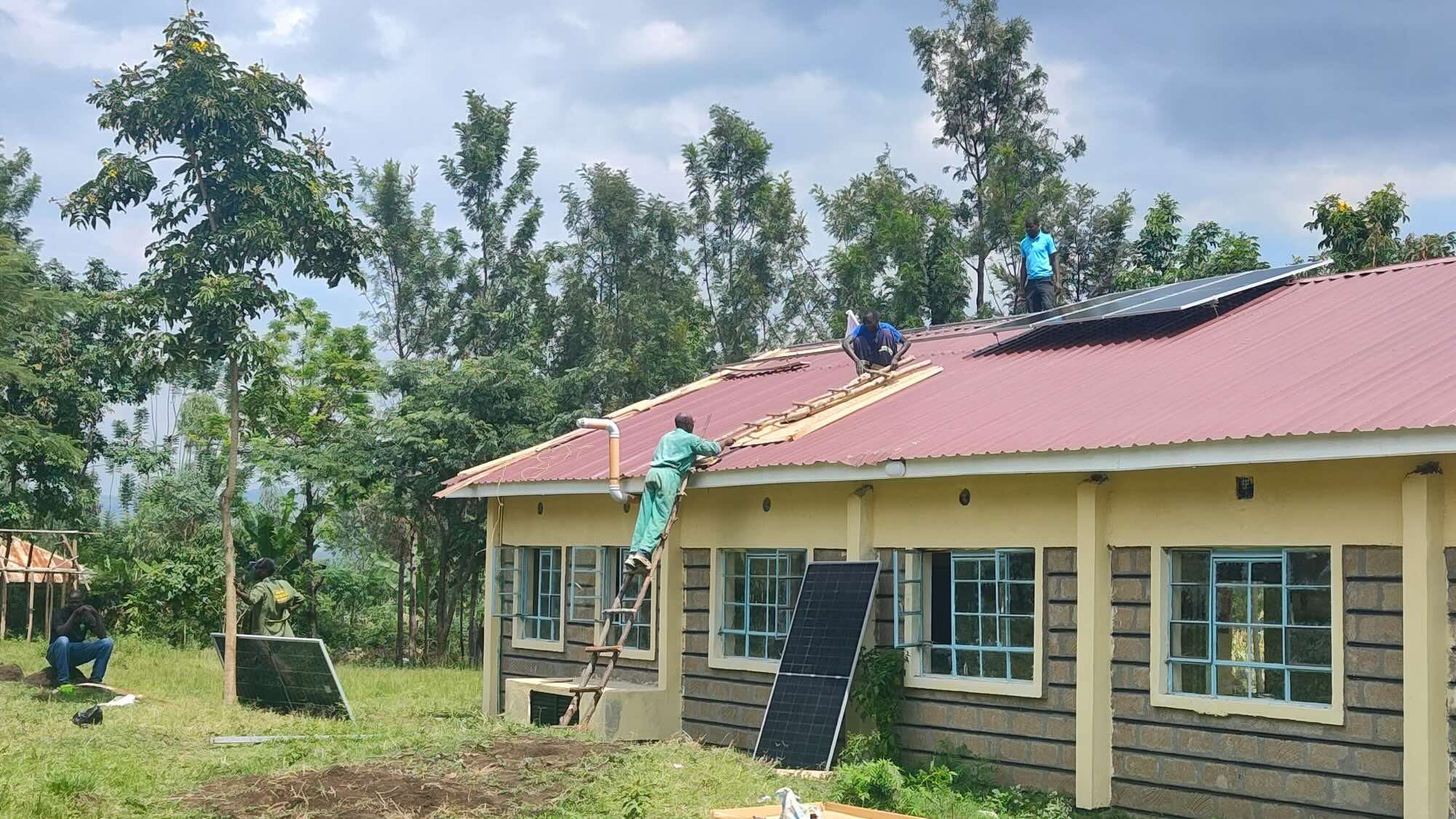 The photo shows the installation of solar panels at a small school in a natural setting.